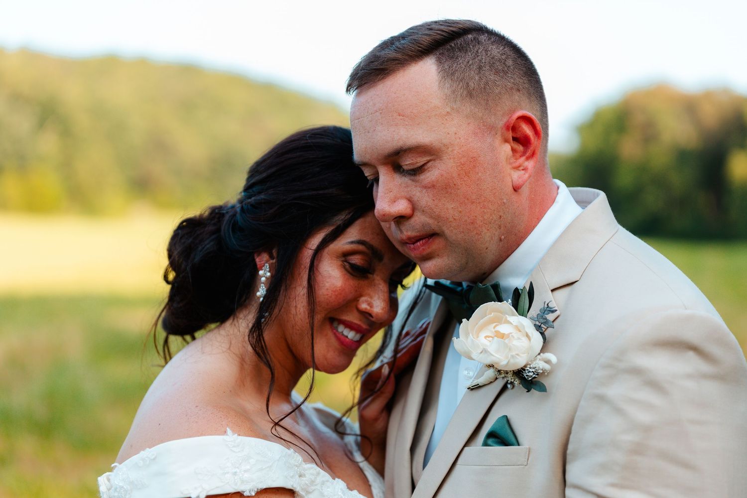 Bride and groom sharing tender moment outdoors, groom in beige suit with boutonniere, bride in white dress with updo hairstyle.