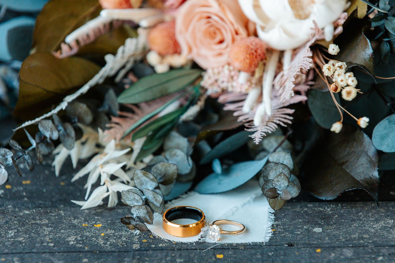 Gold wedding rings resting on fabric beside a romantic bridal bouquet with peach roses and eucalyptus.