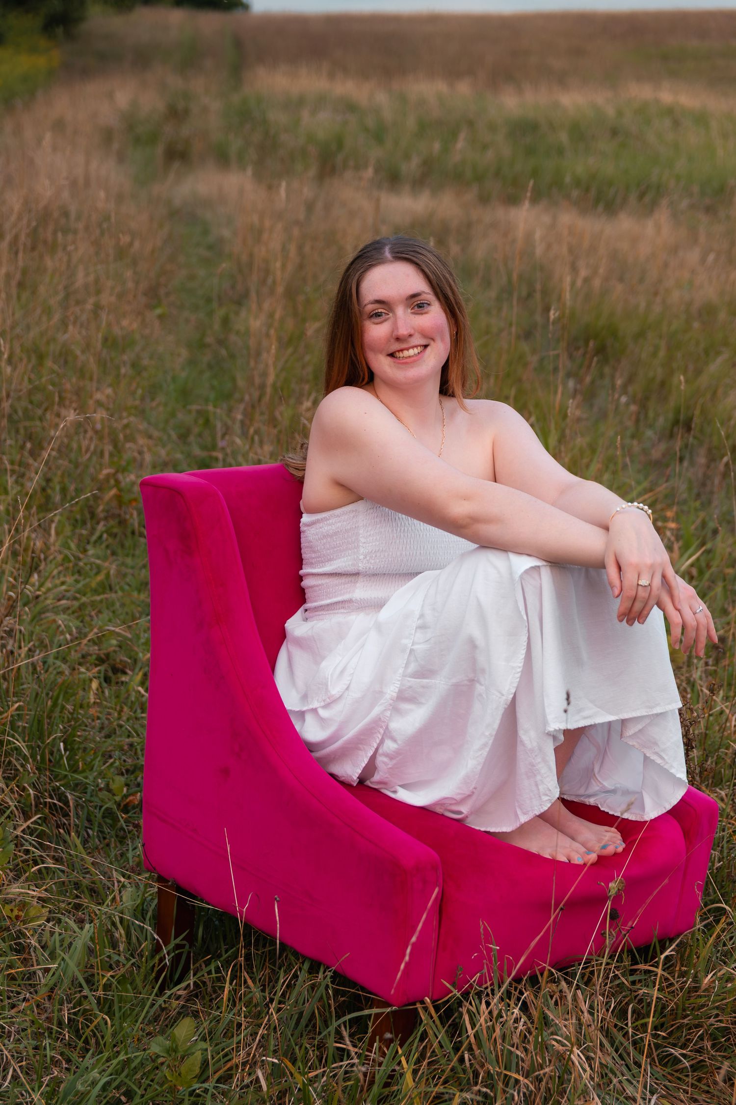 Smiling woman in white outfit sitting barefoot on a pink chair in a grassy field.