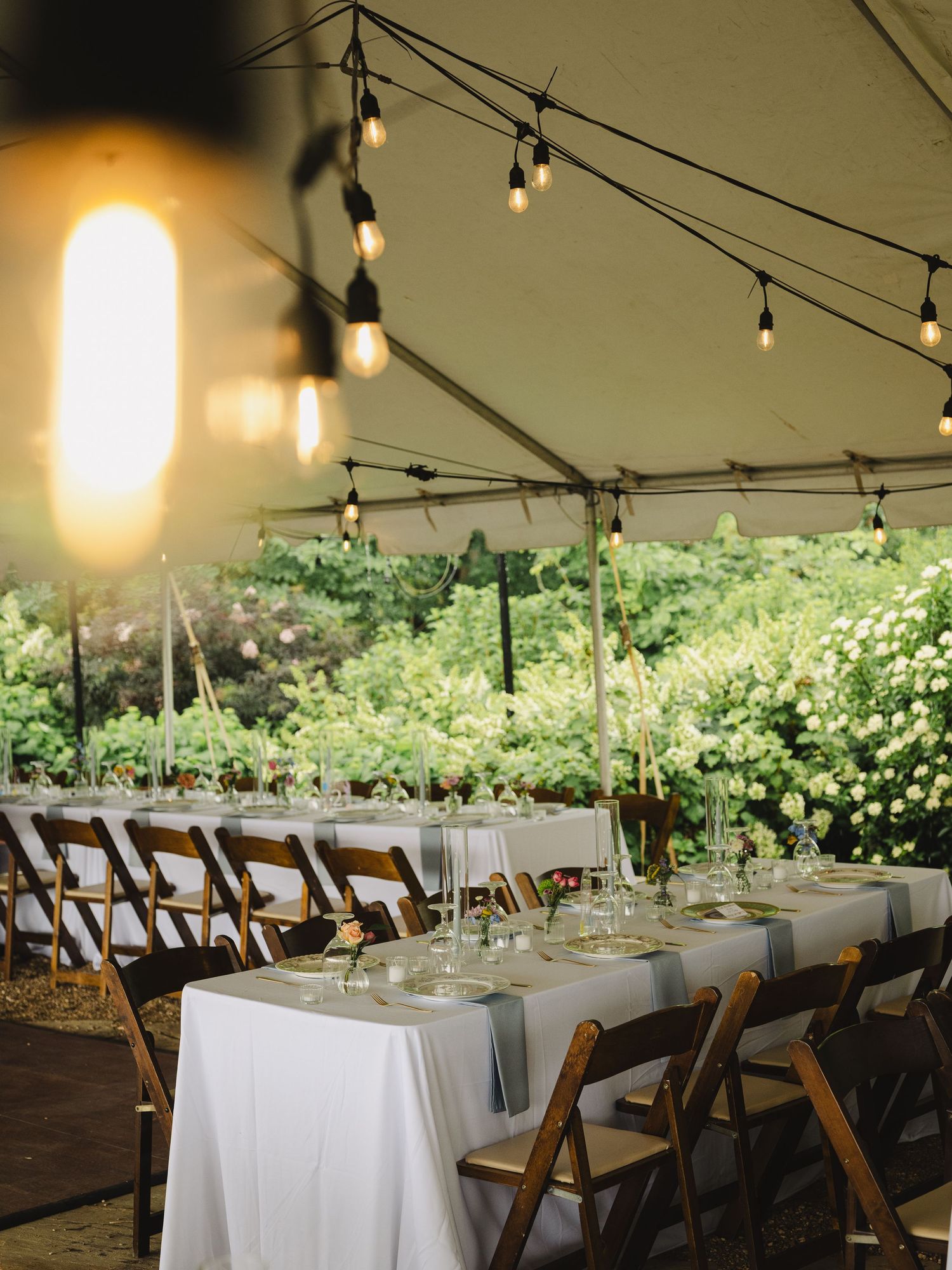 Elegant outdoor wedding reception under a tent with string lights, long white tables, wooden chairs, and lush greenery backdrop.