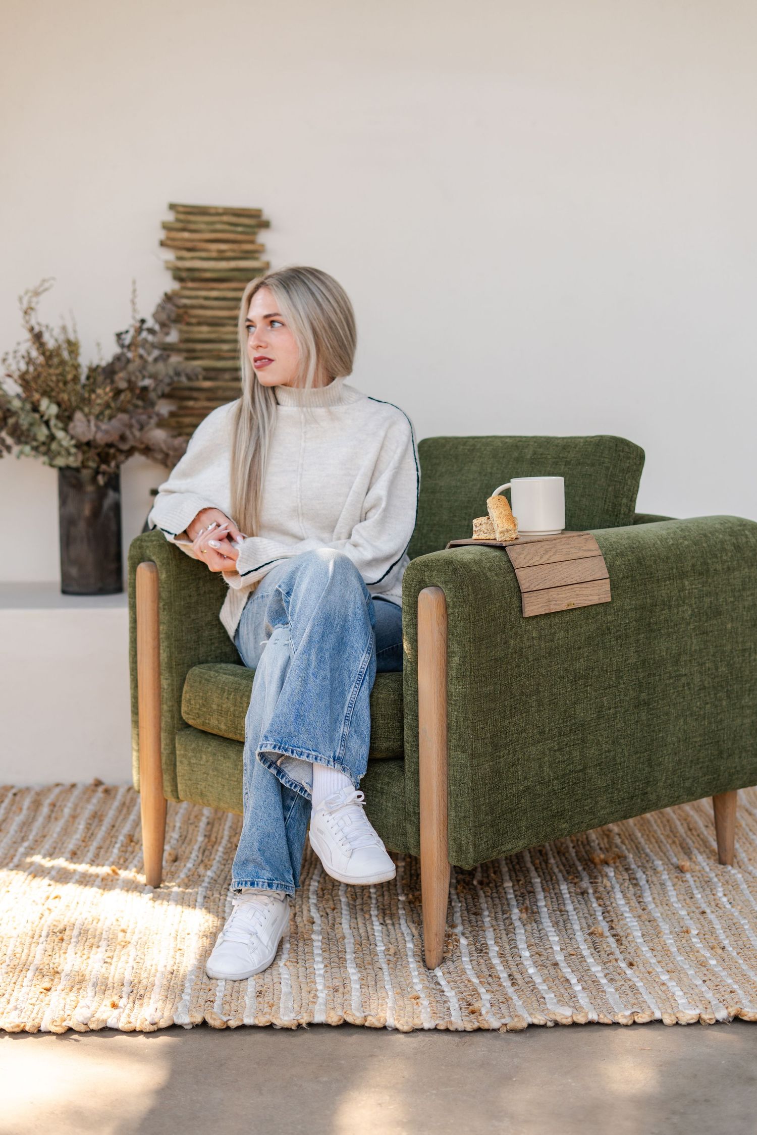 Young woman in white sweater and jeans relaxing in green armchair with coffee mug on wooden tray during a considered product photoshoot.