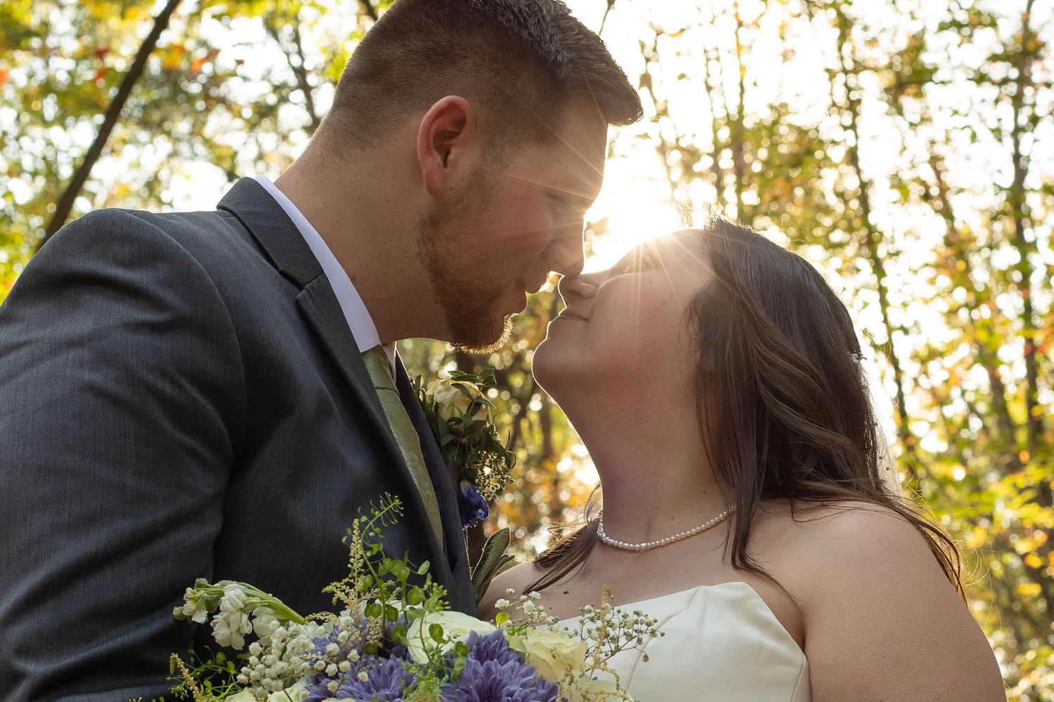 A newlywed couple shares a romantic kiss outdoors, with the bride holding a purple and white bouquet in golden sunlight.