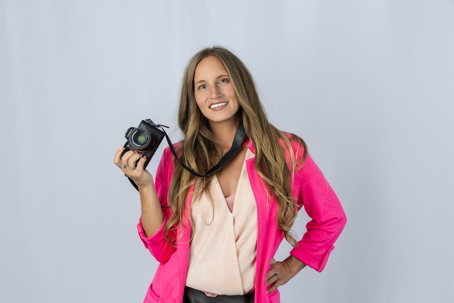 Woman in pink blazer smiling and holding a camera against a light gray background.