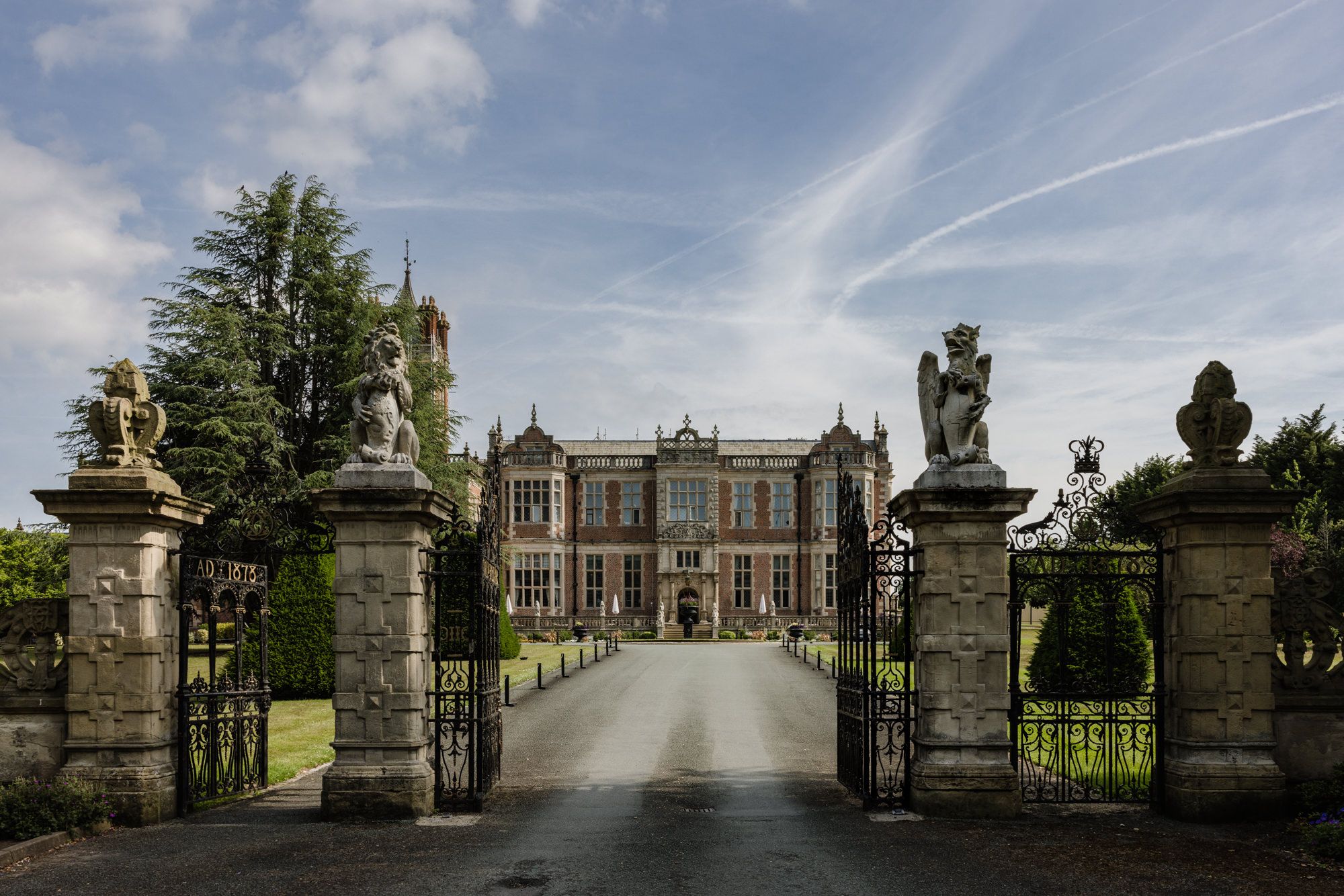 Grand English manor house with ornate iron gates, stone statues, and a tree-lined driveway under a blue sky.