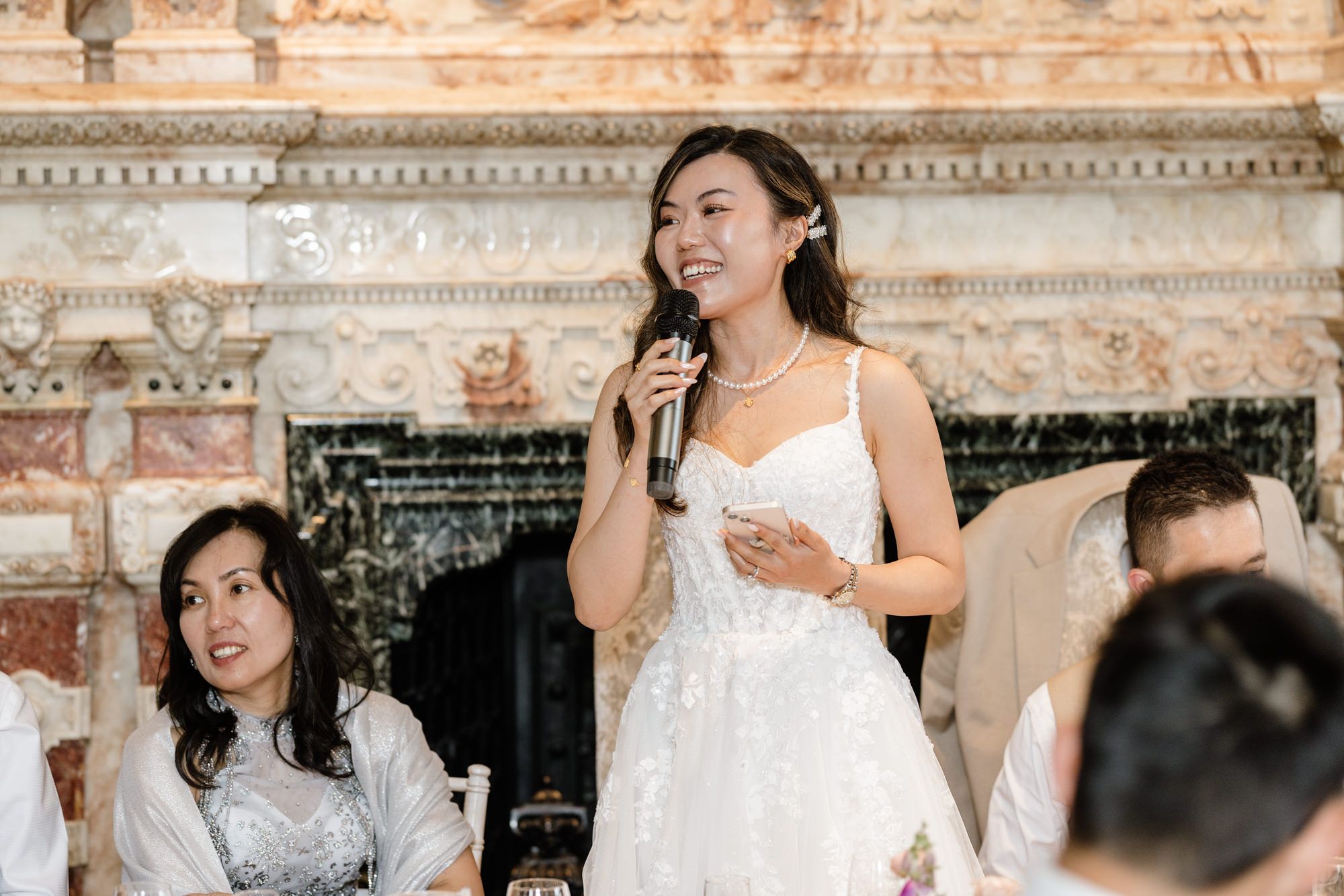 Bride in white lace gown speaking into microphone at wedding reception in elegant venue.