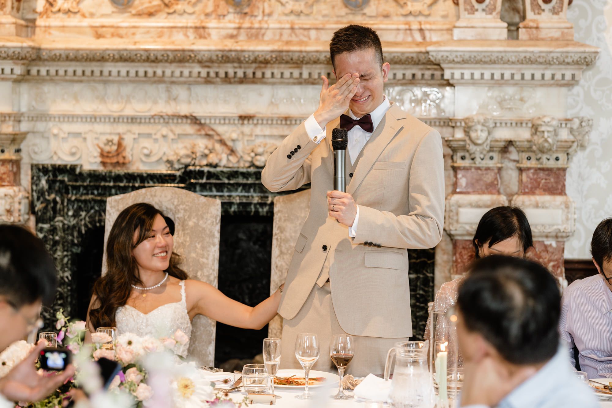 Groom wipes tears while giving wedding speech, bride smiles beside him at elegant reception venue.
