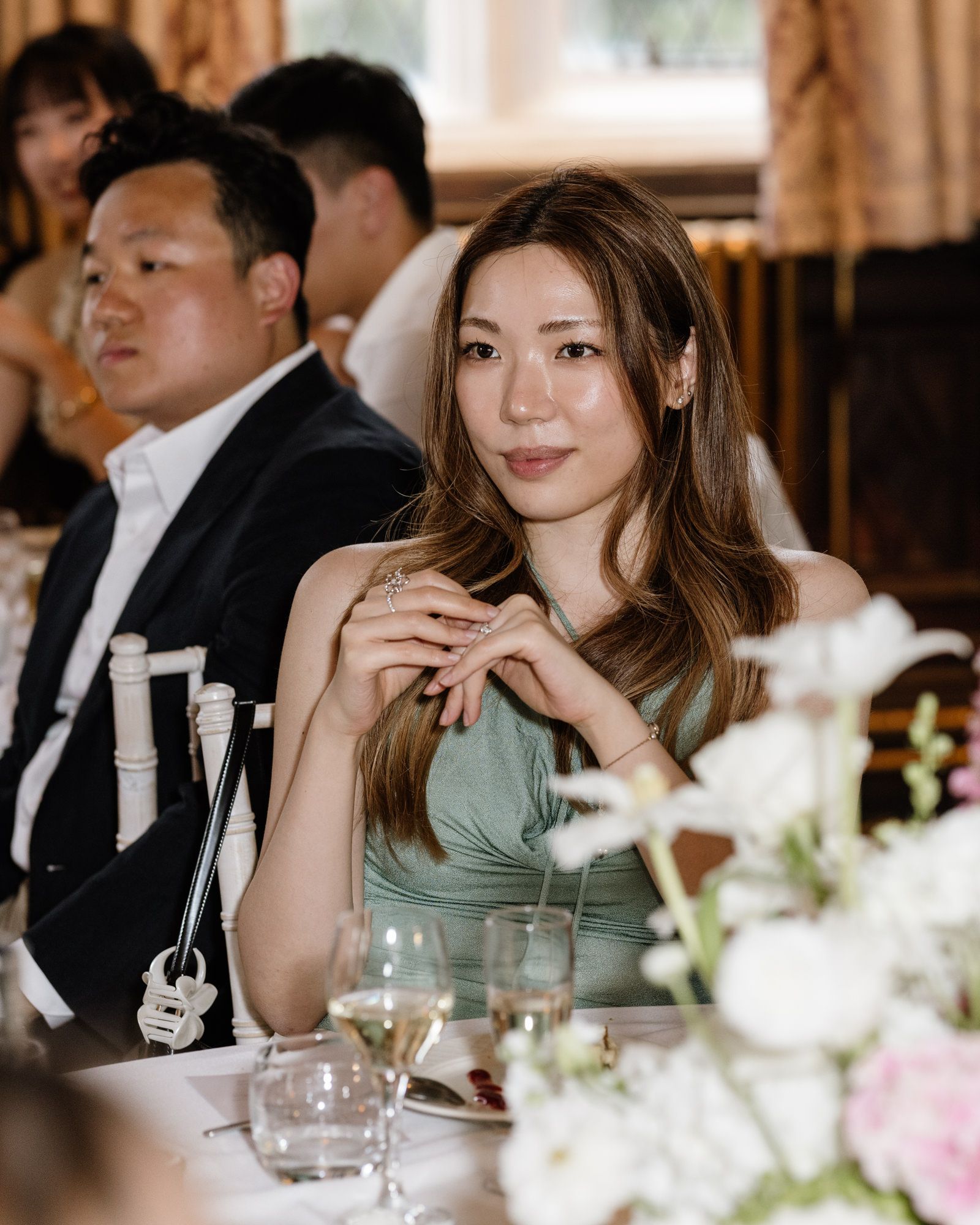 Elegant woman in sage green dress seated at a formal wedding reception table with flowers and wine glasses.