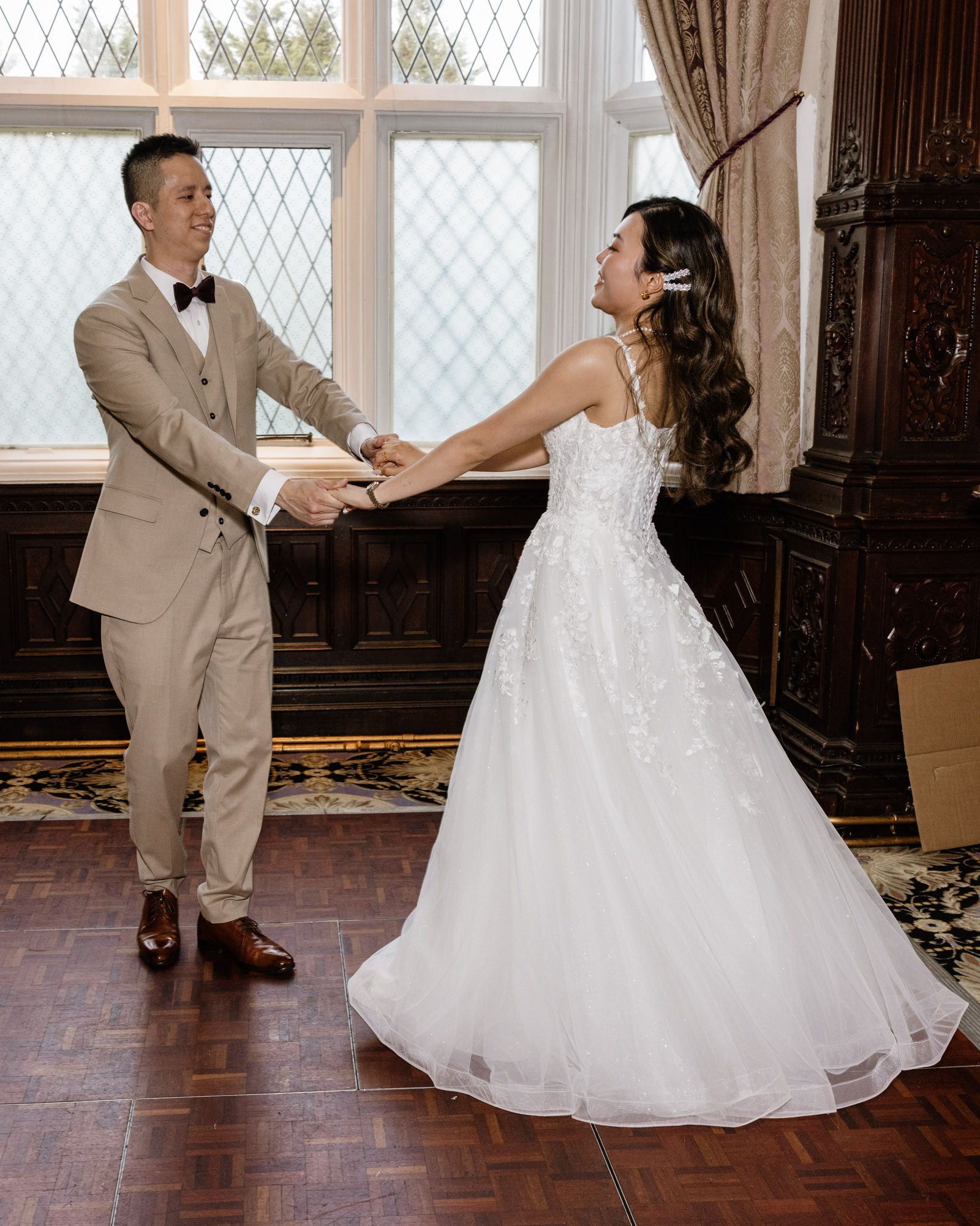 Couple sharing first dance in elegant venue, groom in tan suit and bride in white lace ball gown holding hands.