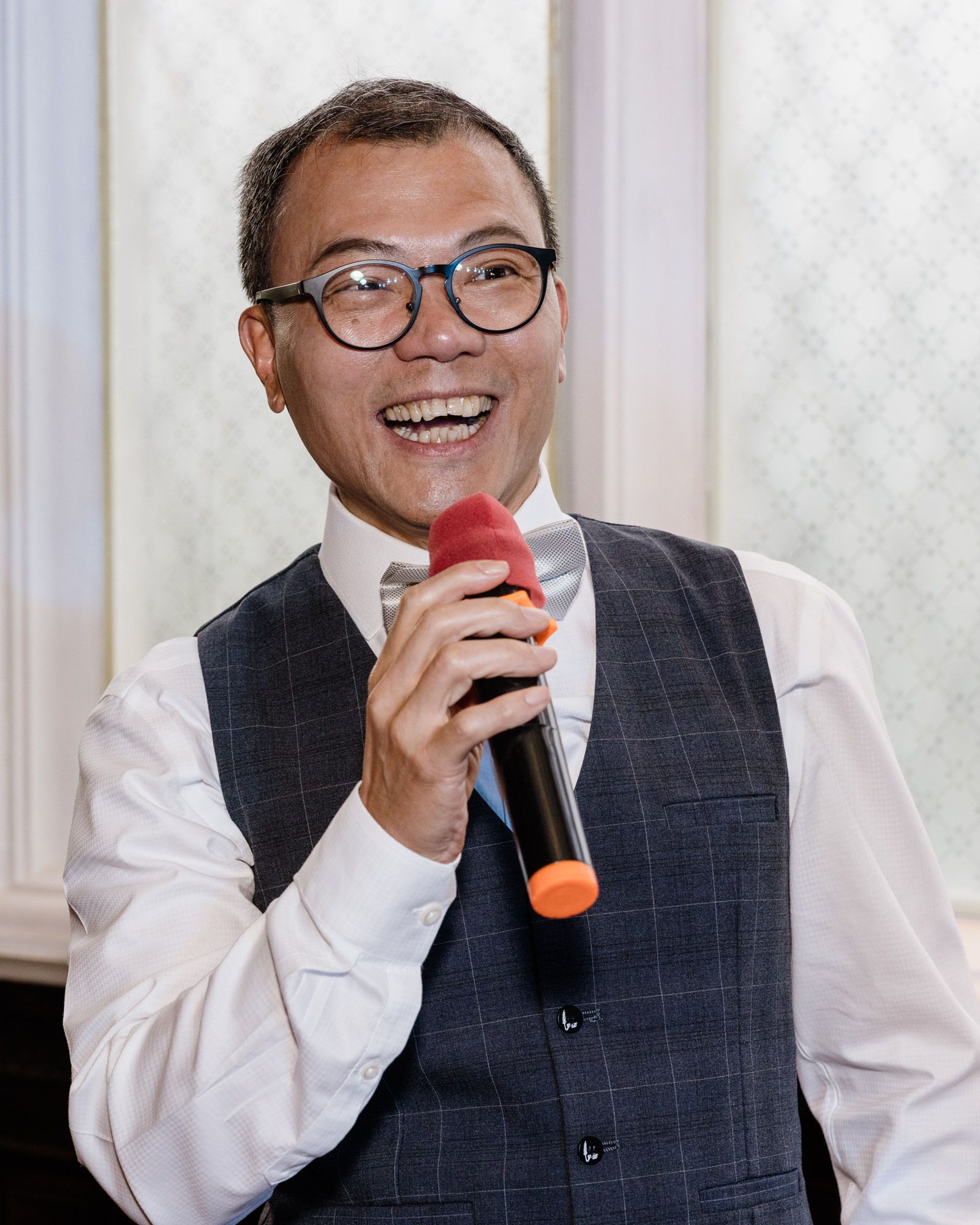 Smiling Asian man in glasses and vest speaking into a red and black microphone at an event.