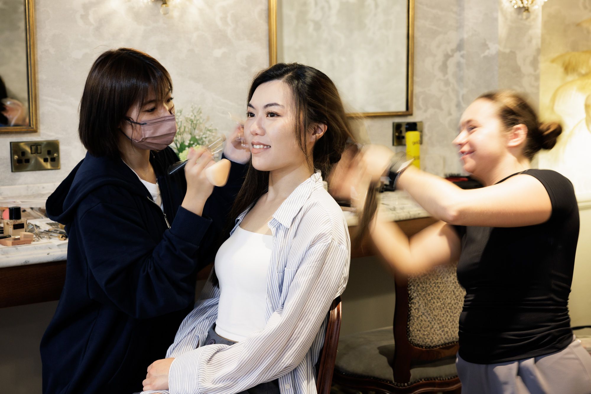 Bride smiling as two makeup artists apply makeup and style hair in a bridal preparation room.