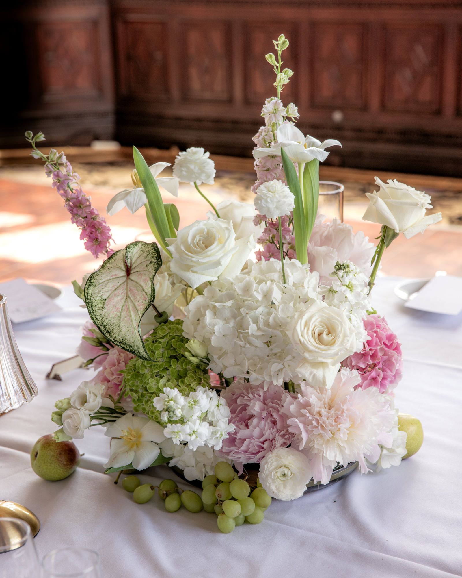 Elegant wedding centerpiece with white roses, pink hydrangeas, green grapes, and mixed blooms on a white tablecloth.