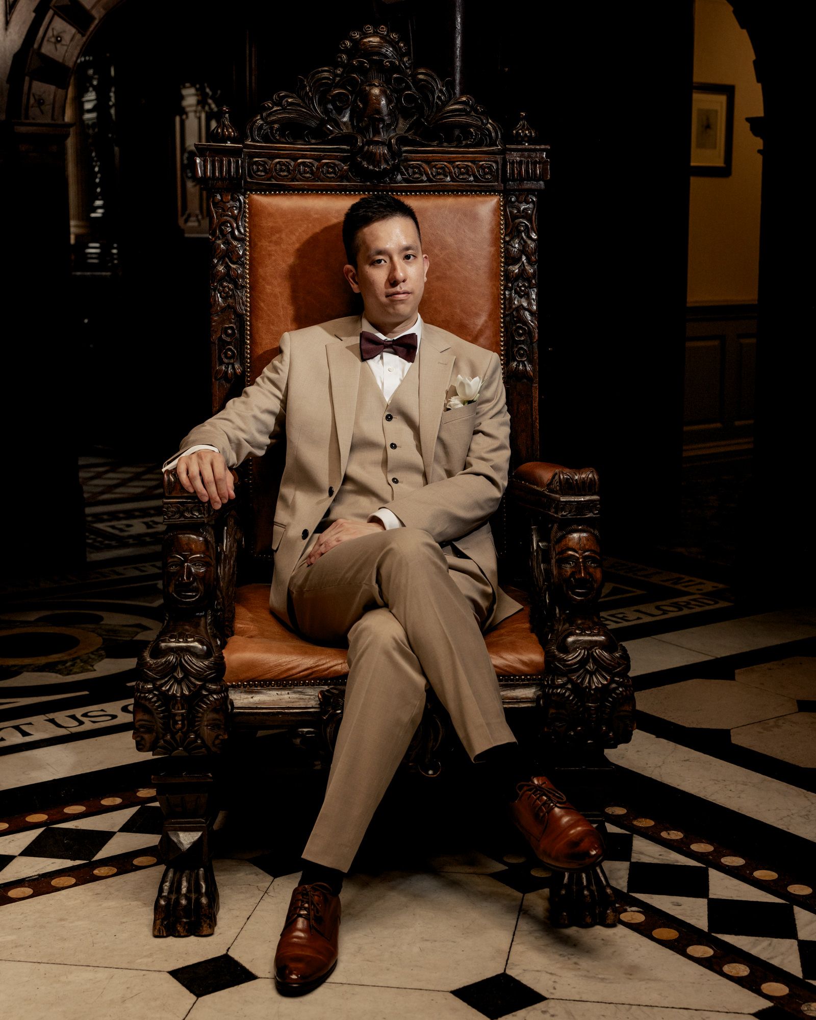 Groom in tan three-piece suit and bow tie poses in ornate leather throne chair.