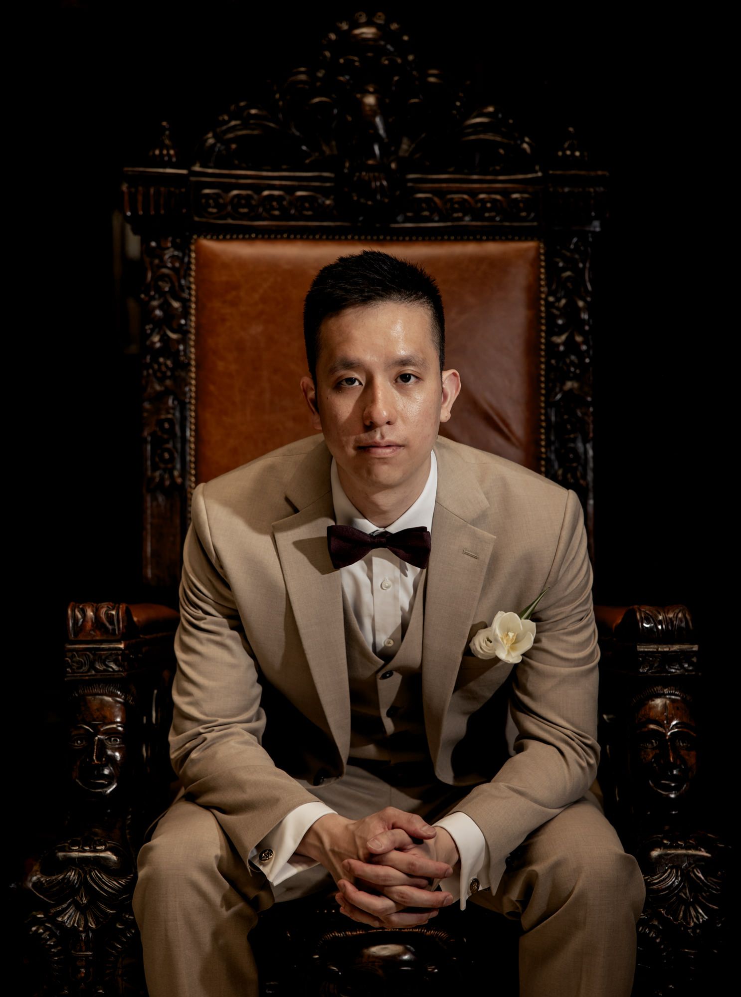 Man in tan suit and bow tie sits in ornate throne chair with clasped hands.