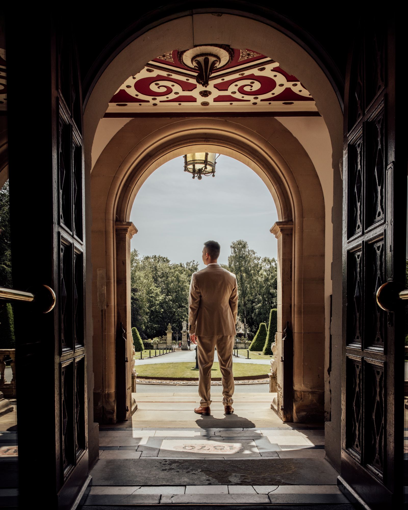 Man in suit standing in ornate arched doorway overlooking formal garden landscape.