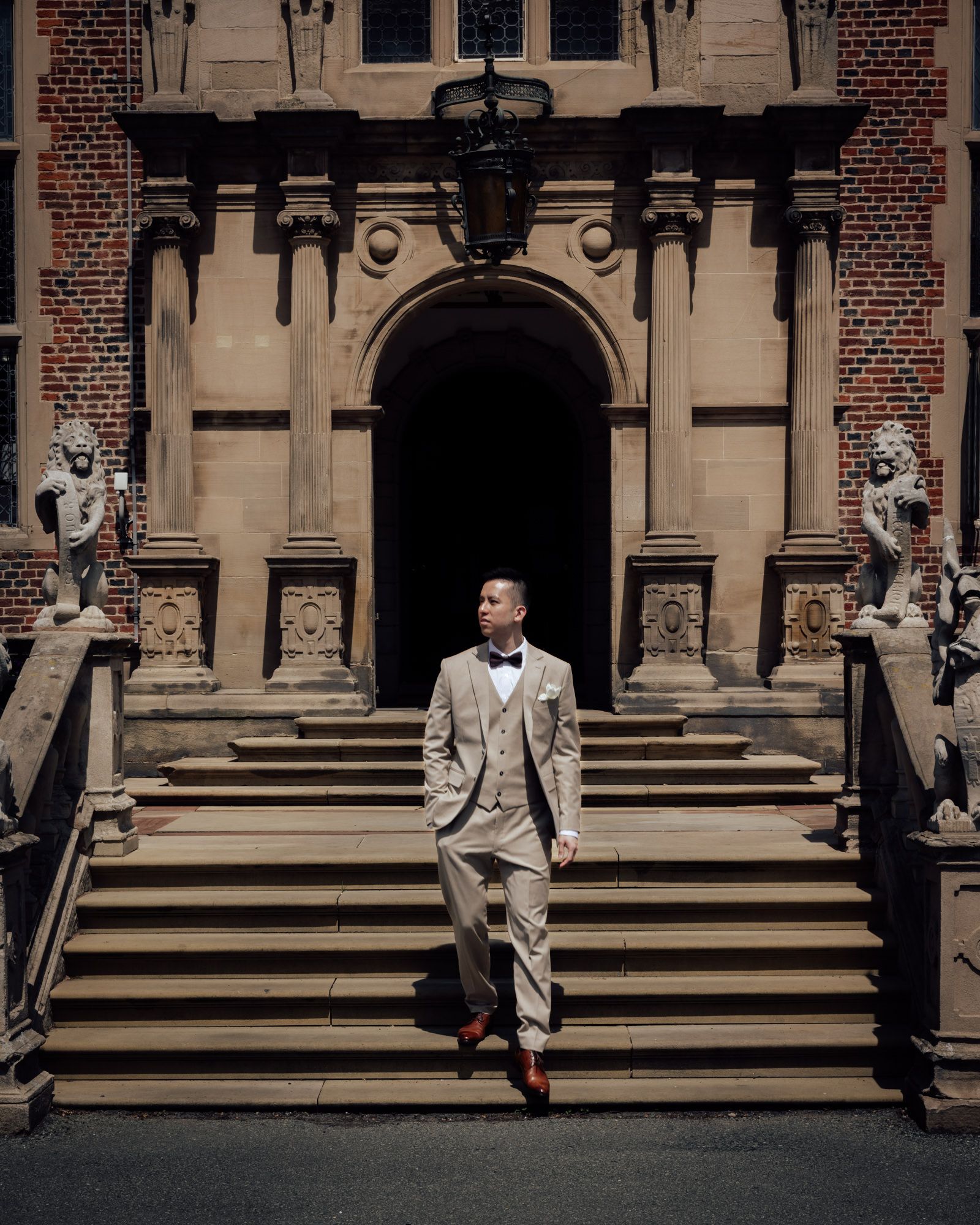 A well-dressed man in a tan suit and bow tie stands on stone steps of an elegant brick building.