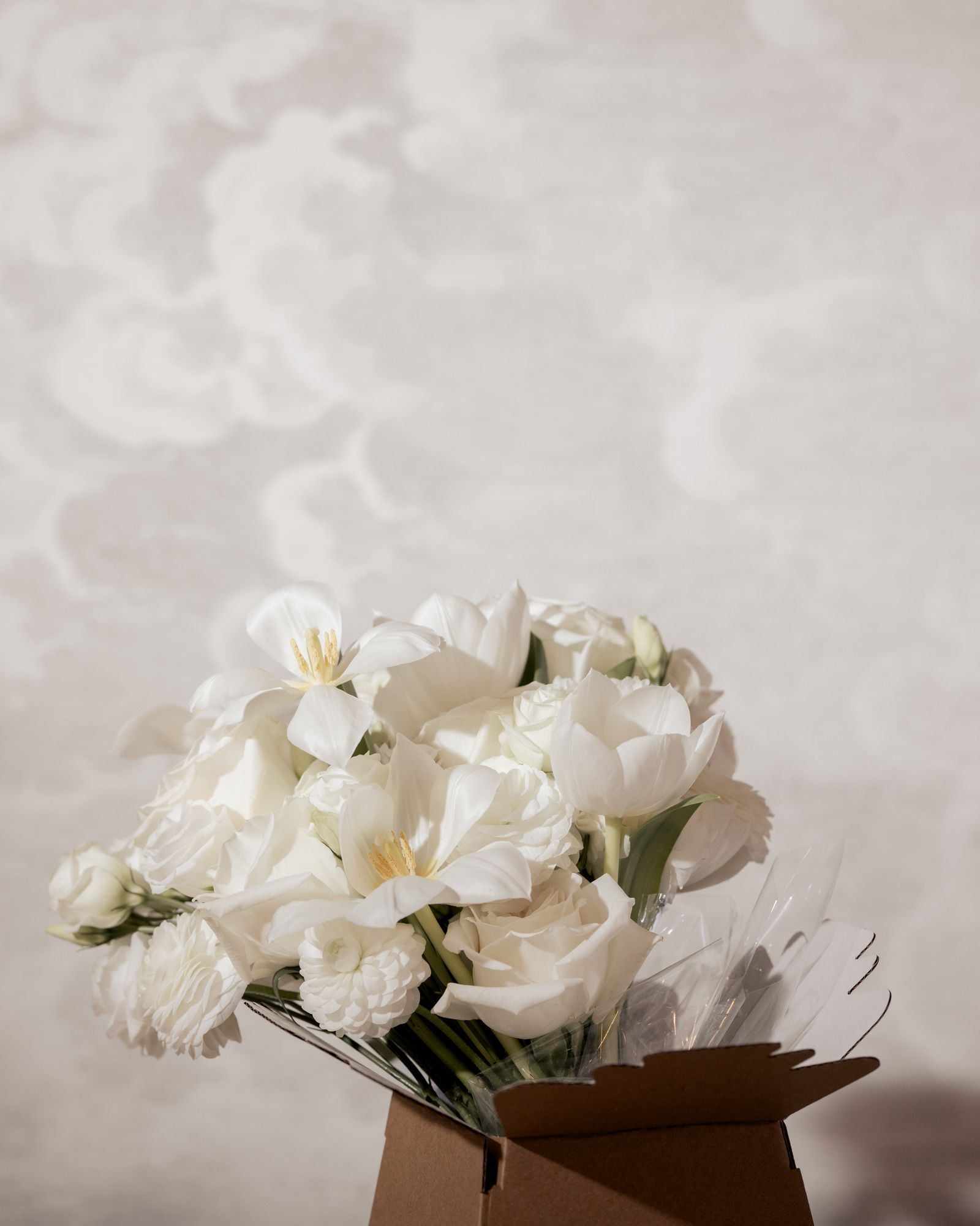 Elegant bouquet of white tulips and roses arranged in a kraft paper box against a soft gray background.