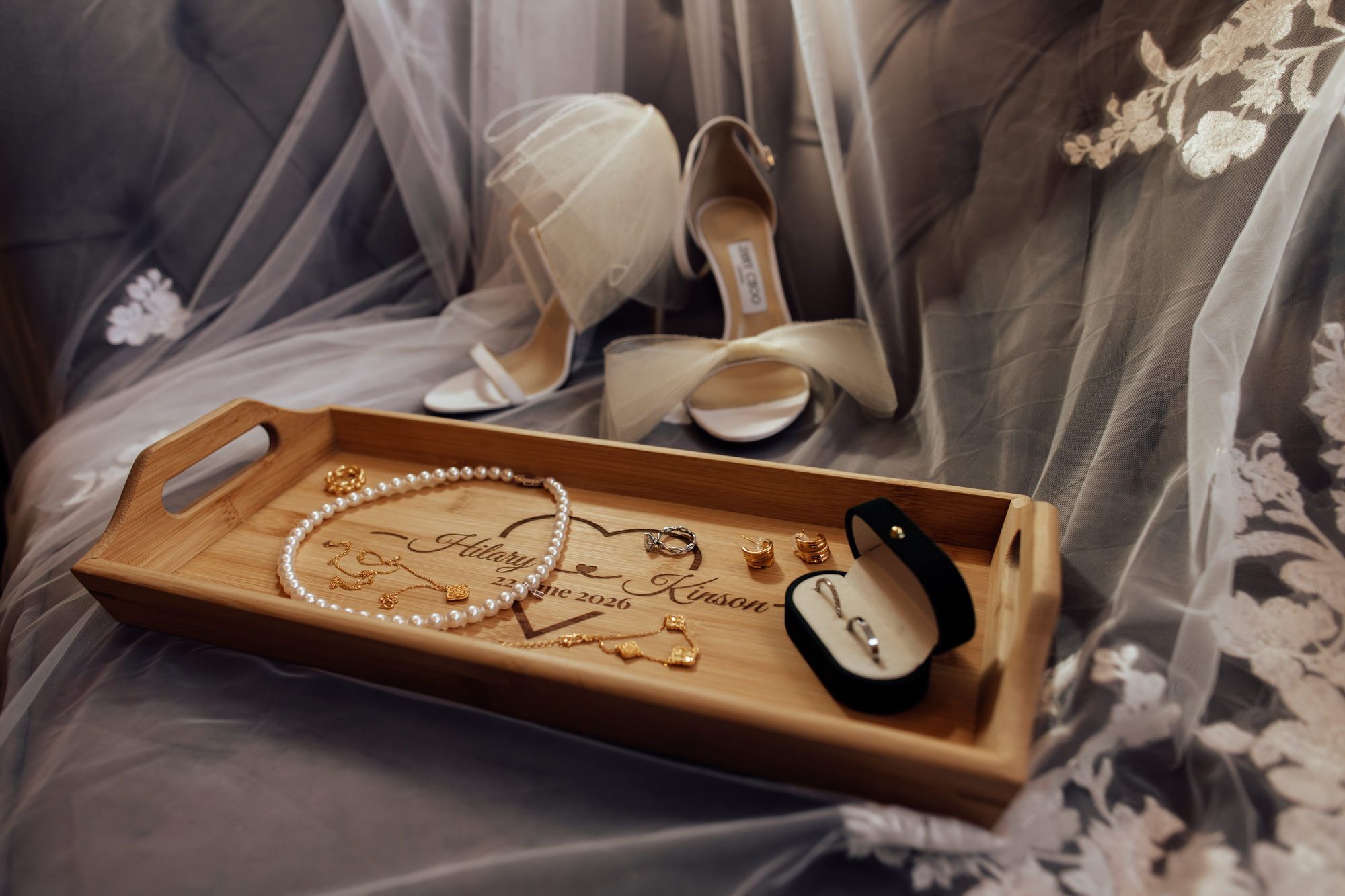Elegant bridal flat lay with pearl necklace, gold jewelry, heels, and engraved wooden tray on lace veil.