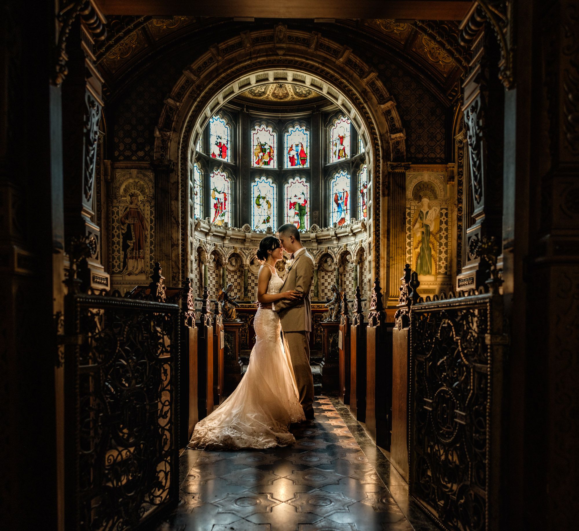 A bride and groom share a romantic embrace in an ornate church with stunning stained glass windows behind them.