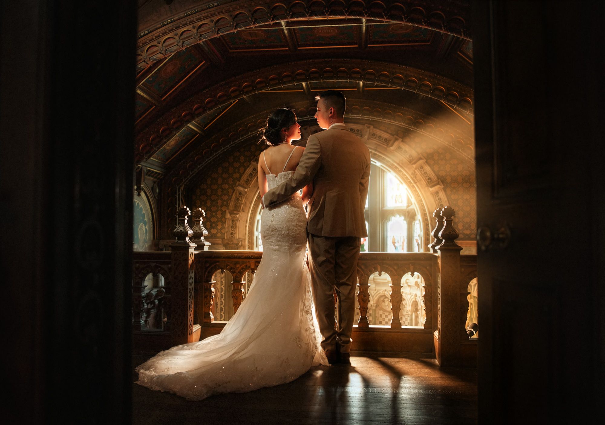 Bride and groom share romantic moment in golden light inside ornate castle hall.
