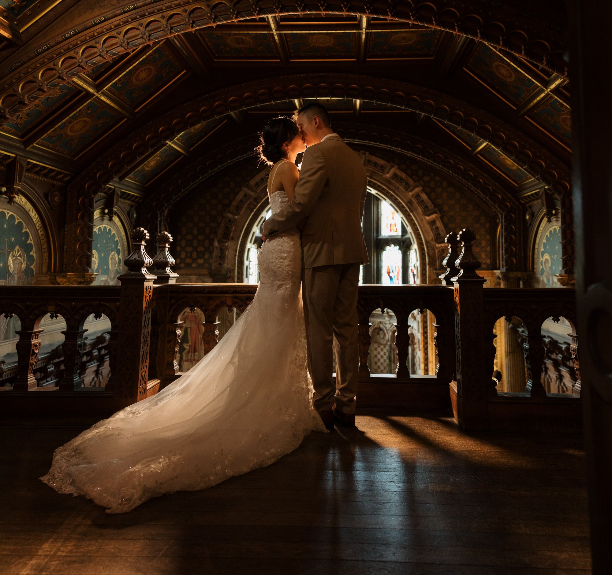 Bride and groom share romantic embrace in ornate Gothic chapel with arched windows and warm sunlight.
