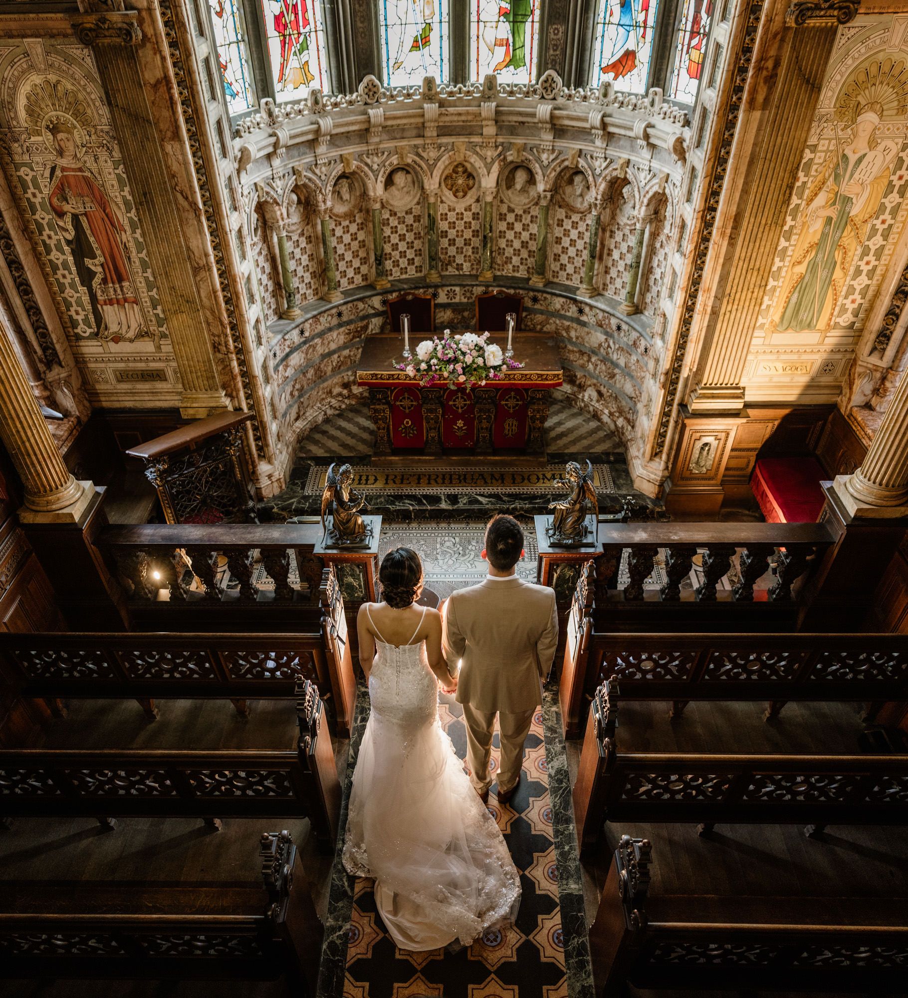 Bride and groom stand at ornate church altar with stunning stained glass windows and intricate golden ceiling details.