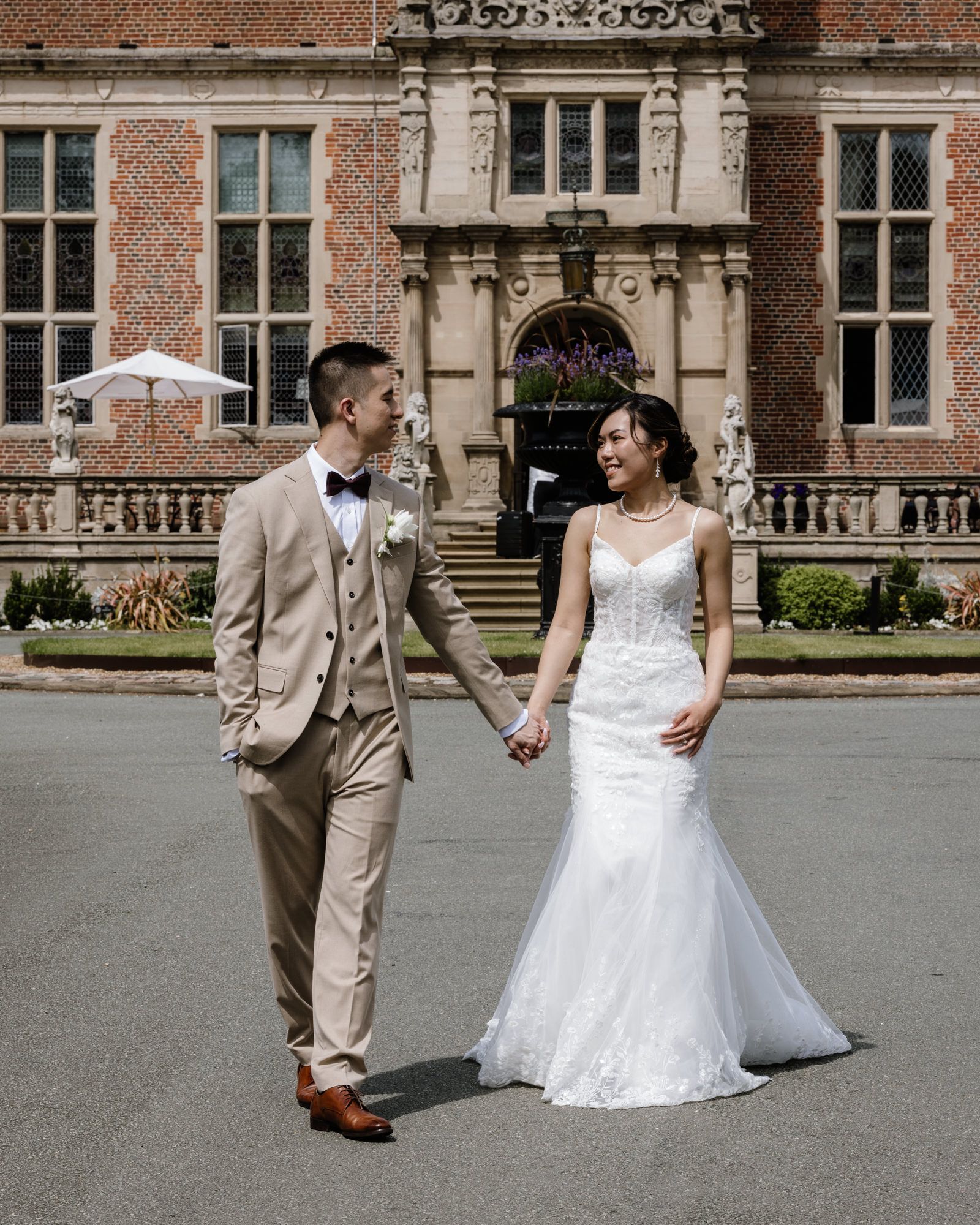 Bride and groom holding hands in front of elegant brick manor on their wedding day.