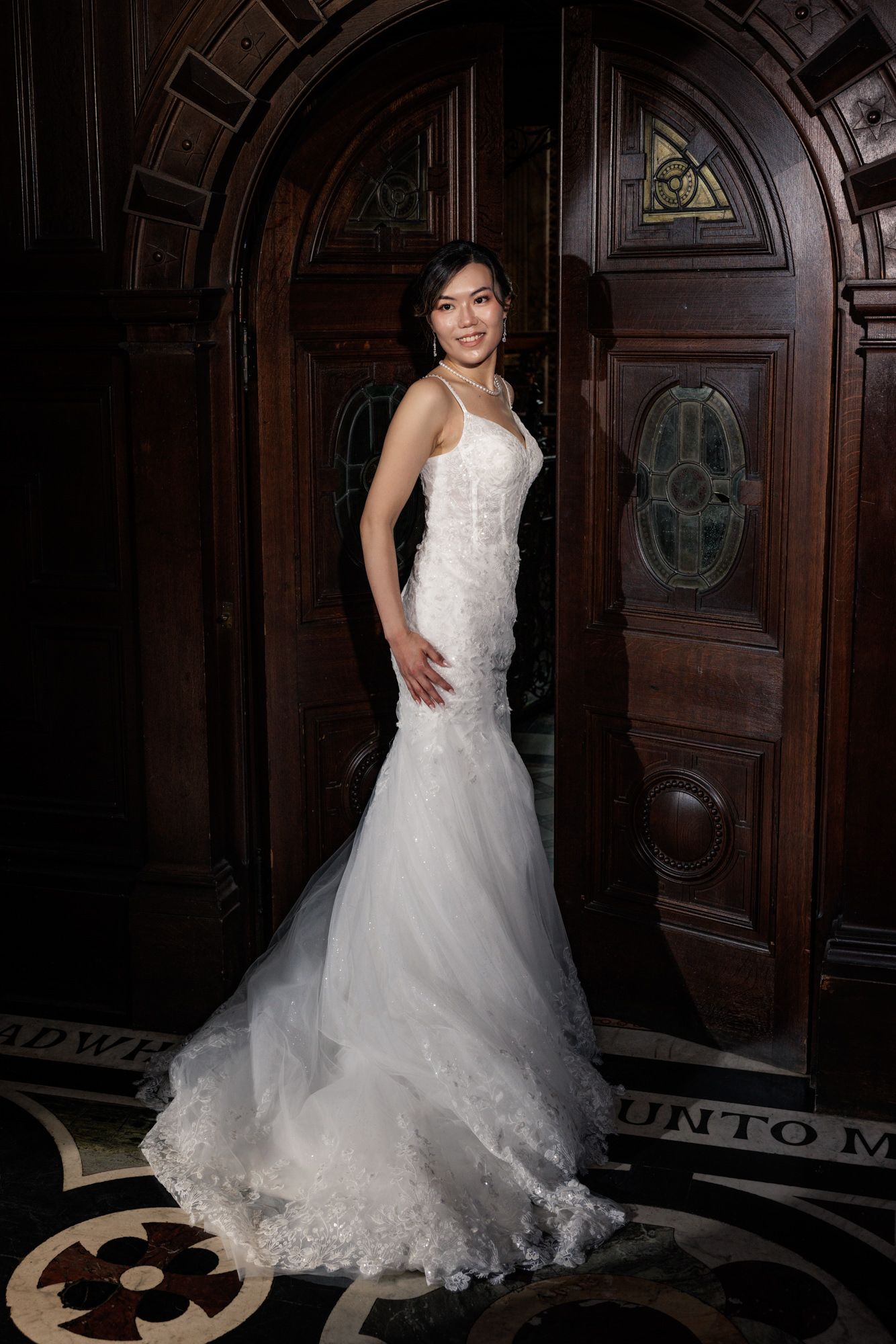 Bride in a white lace mermaid wedding gown posing elegantly in front of ornate wooden doors.