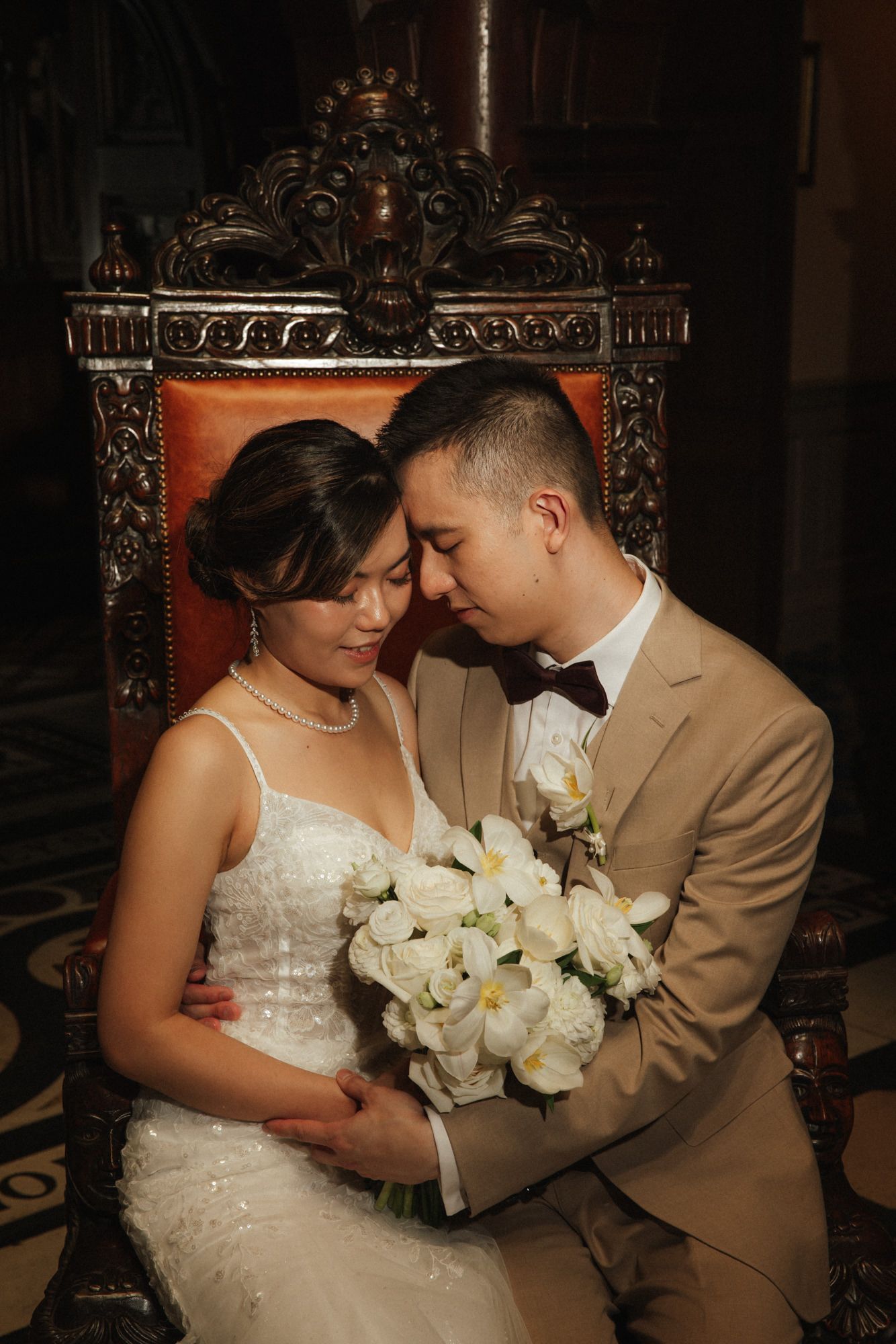 Asian bride and groom in elegant wedding attire share an intimate moment seated in an ornate chair with white floral bouquet.