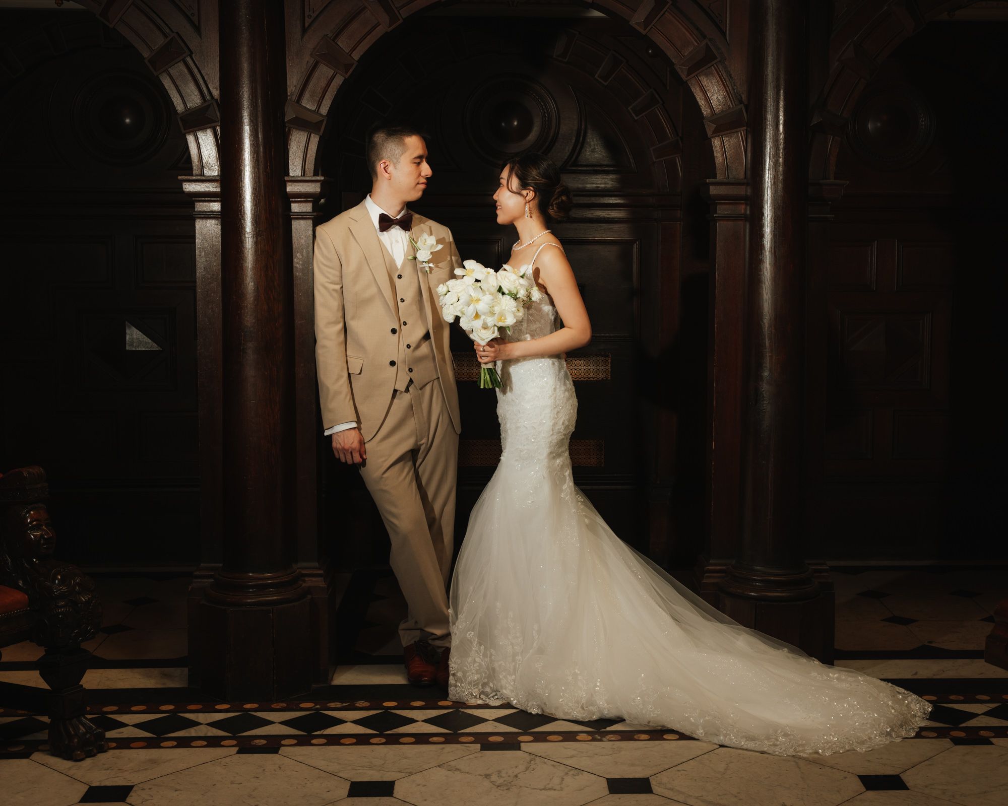 Bride in lace mermaid gown and groom in tan tuxedo share a romantic moment in an elegant dark wood foyer.