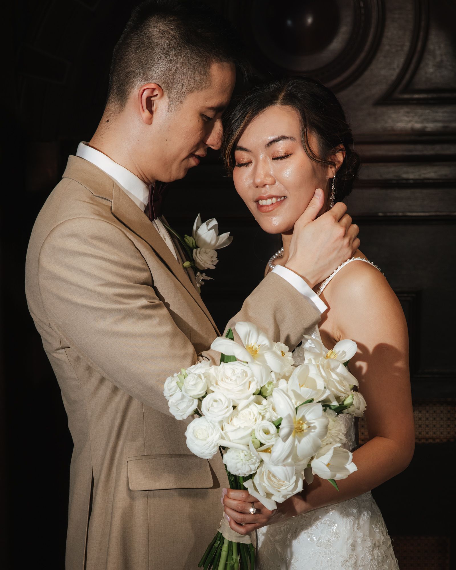 A bride and groom share a tender moment, she holds white roses bouquet, wearing lace dress, he in tan suit.