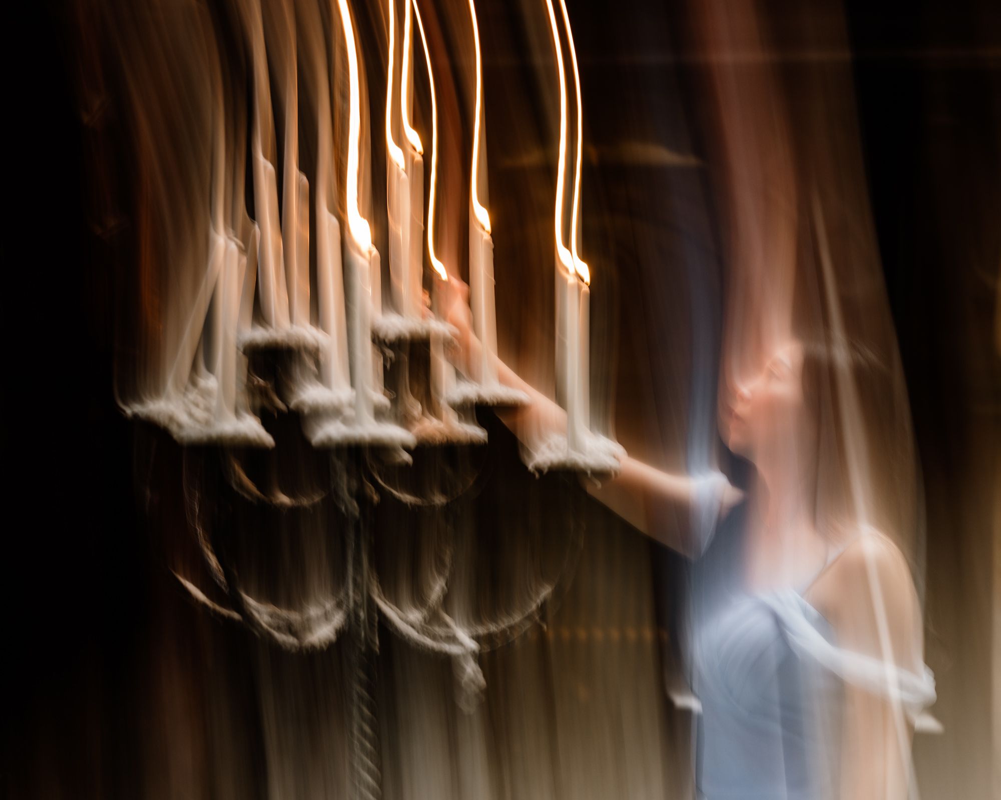 Blurred motion photo of a woman in blue dress lighting candles on an ornate chandelier in dim light.