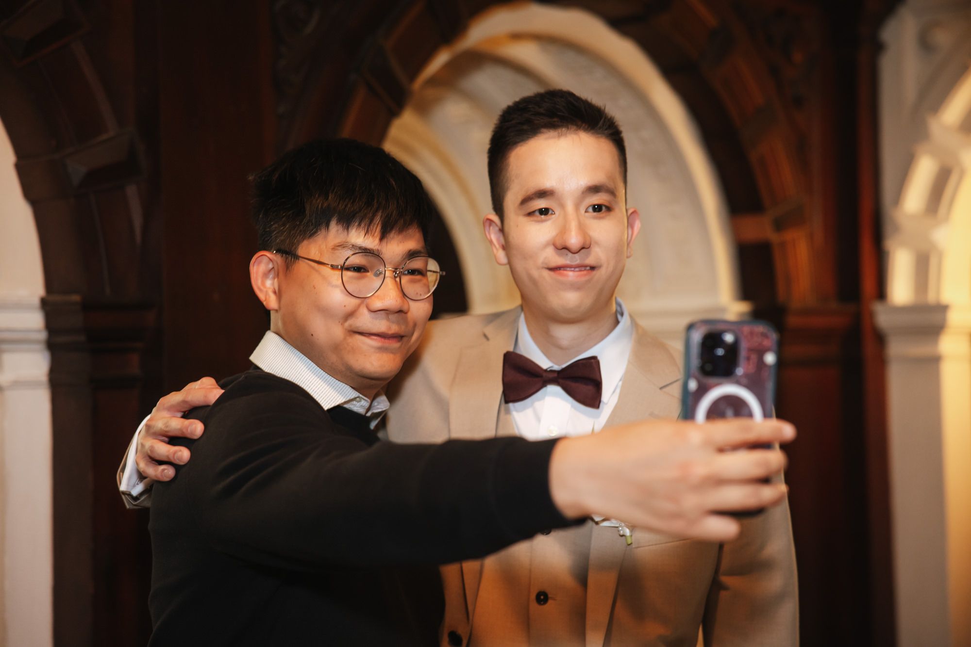 Two men taking a selfie together at an indoor event, one wearing glasses and the other in a tan suit with bow tie.