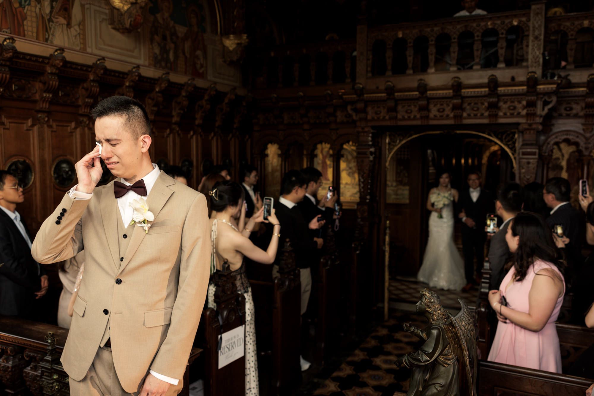 Emotional groom wiping tears in elegant church as bride walks down aisle during wedding ceremony.