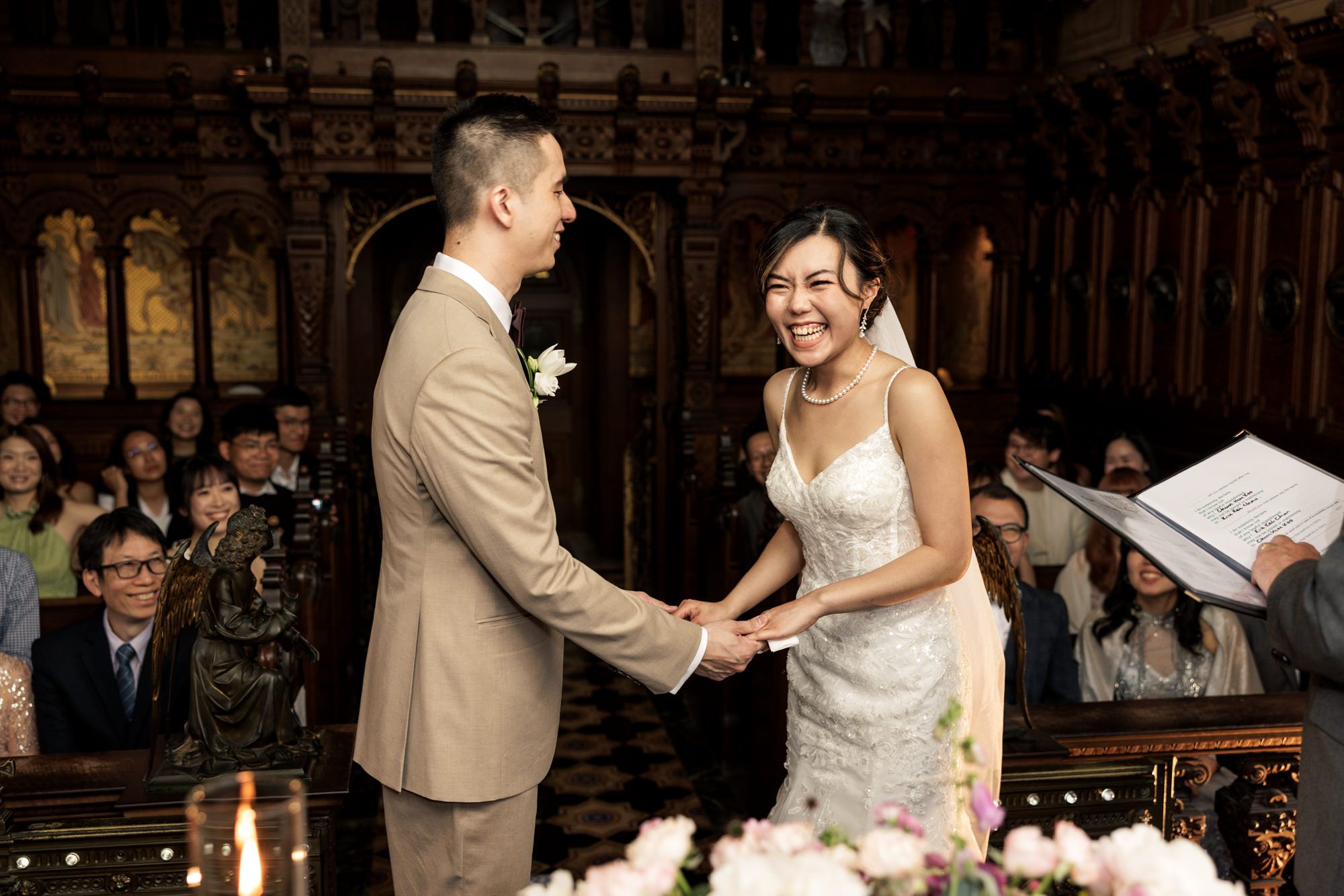Couple holds hands and smiles during wedding ceremony in ornate chapel with guests seated behind them.