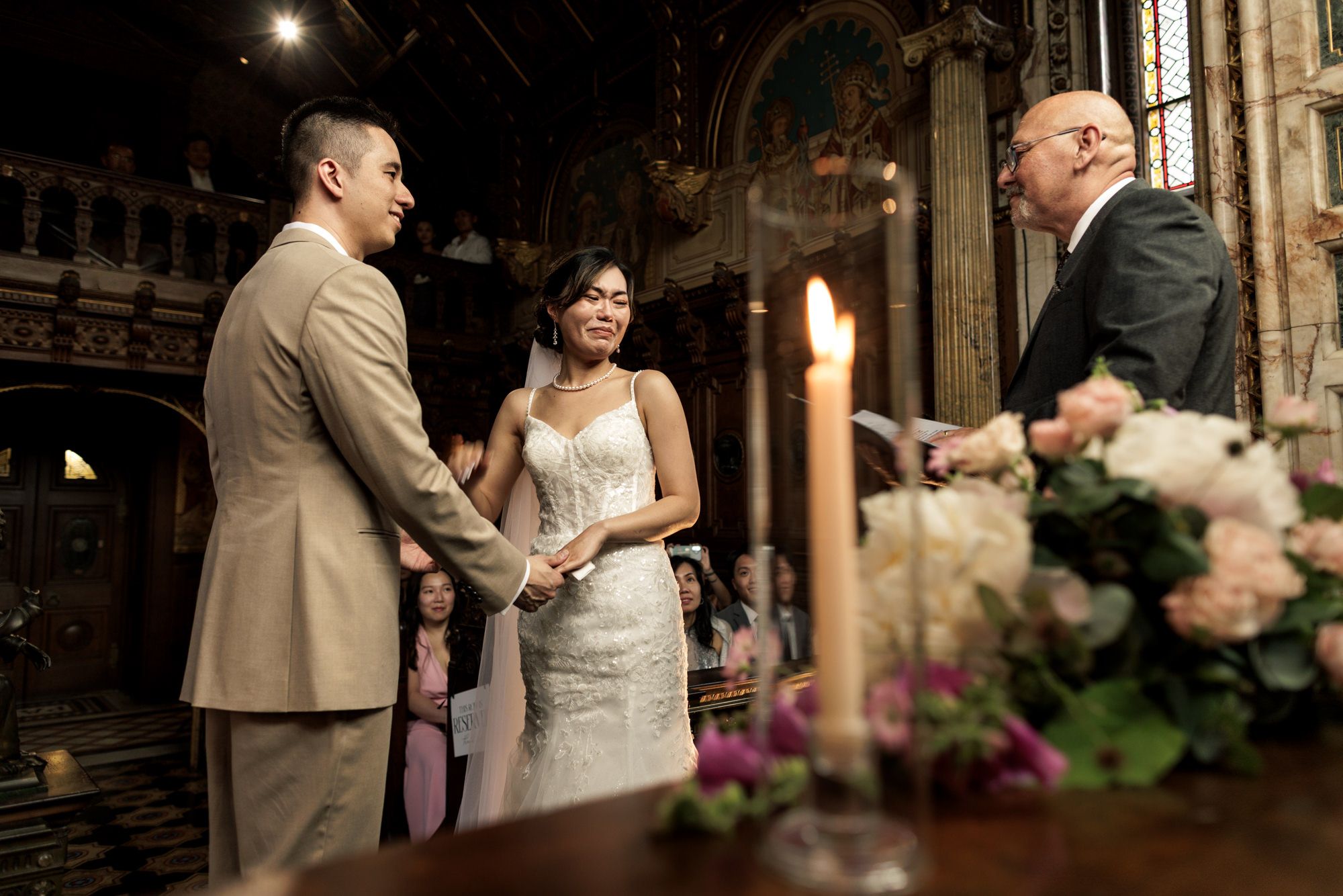 Couple exchanges vows holding hands before officiant at candlelit church wedding altar with floral arrangements.