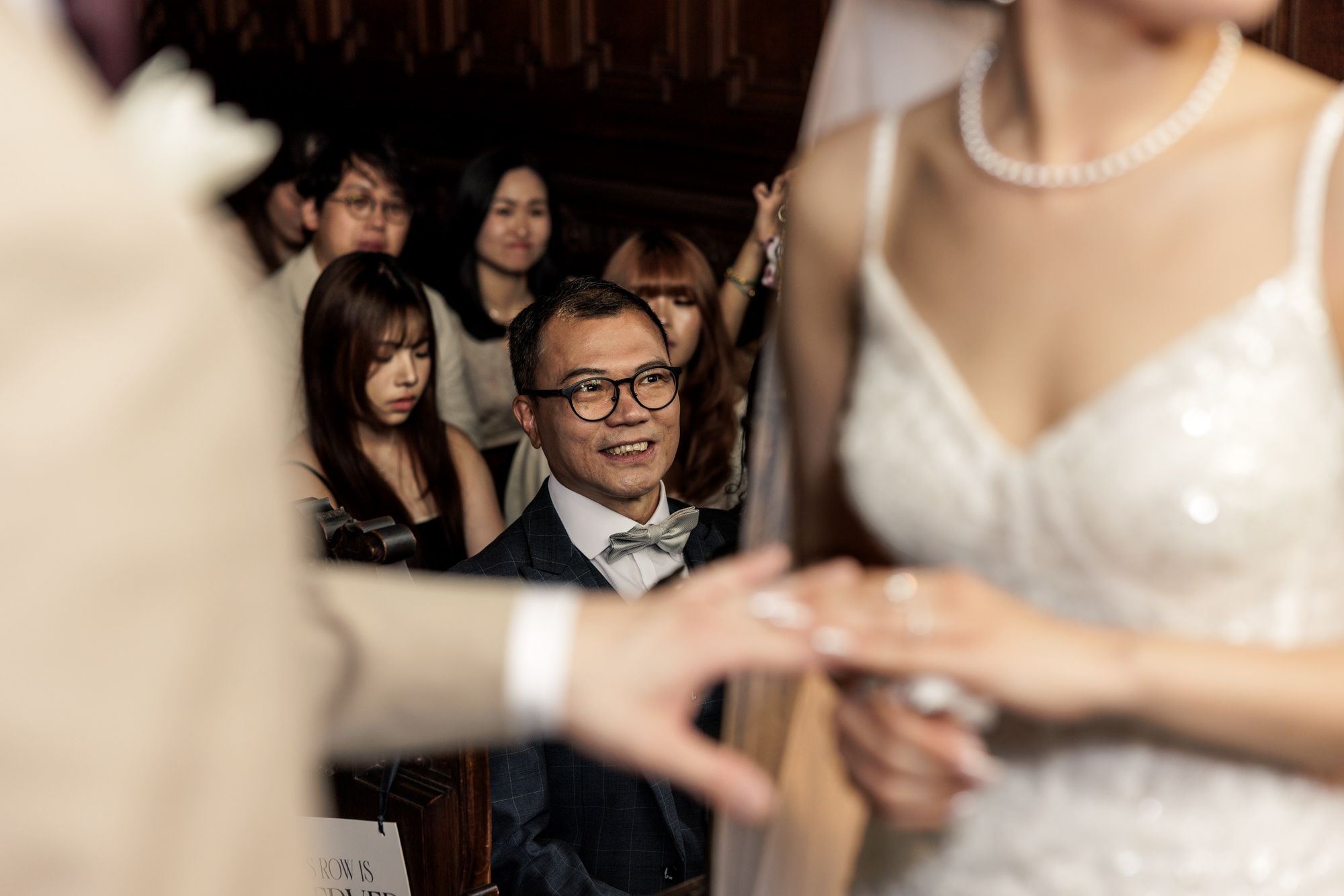 A smiling wedding guest in glasses watches a bride and groom exchange rings during a wedding ceremony.