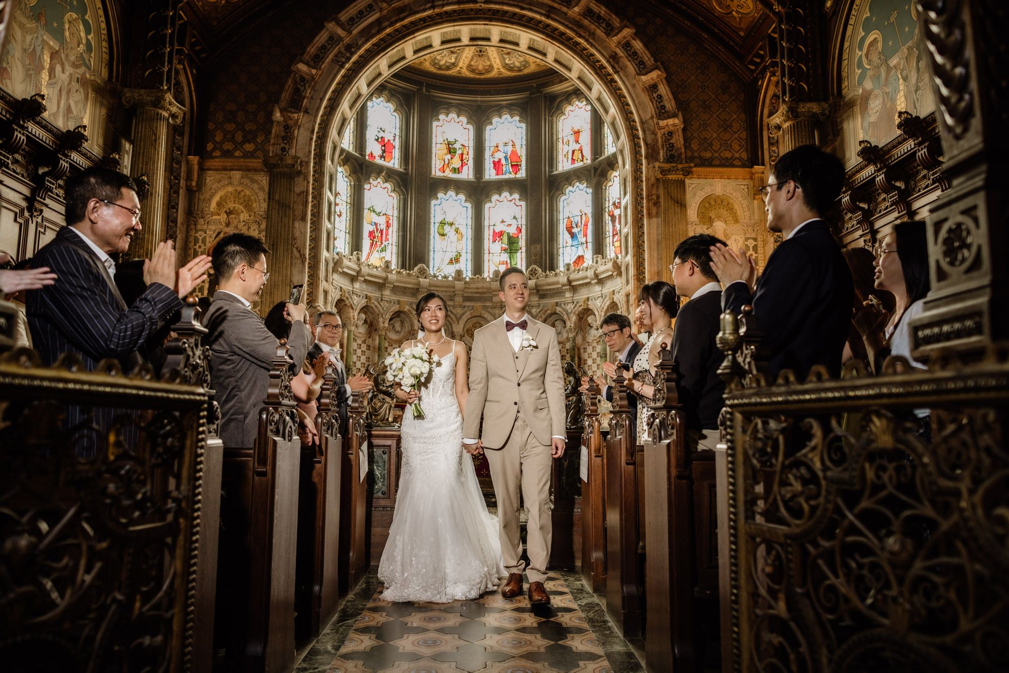 Bride and groom walk down the aisle of an ornate church with stained glass windows at their wedding ceremony.