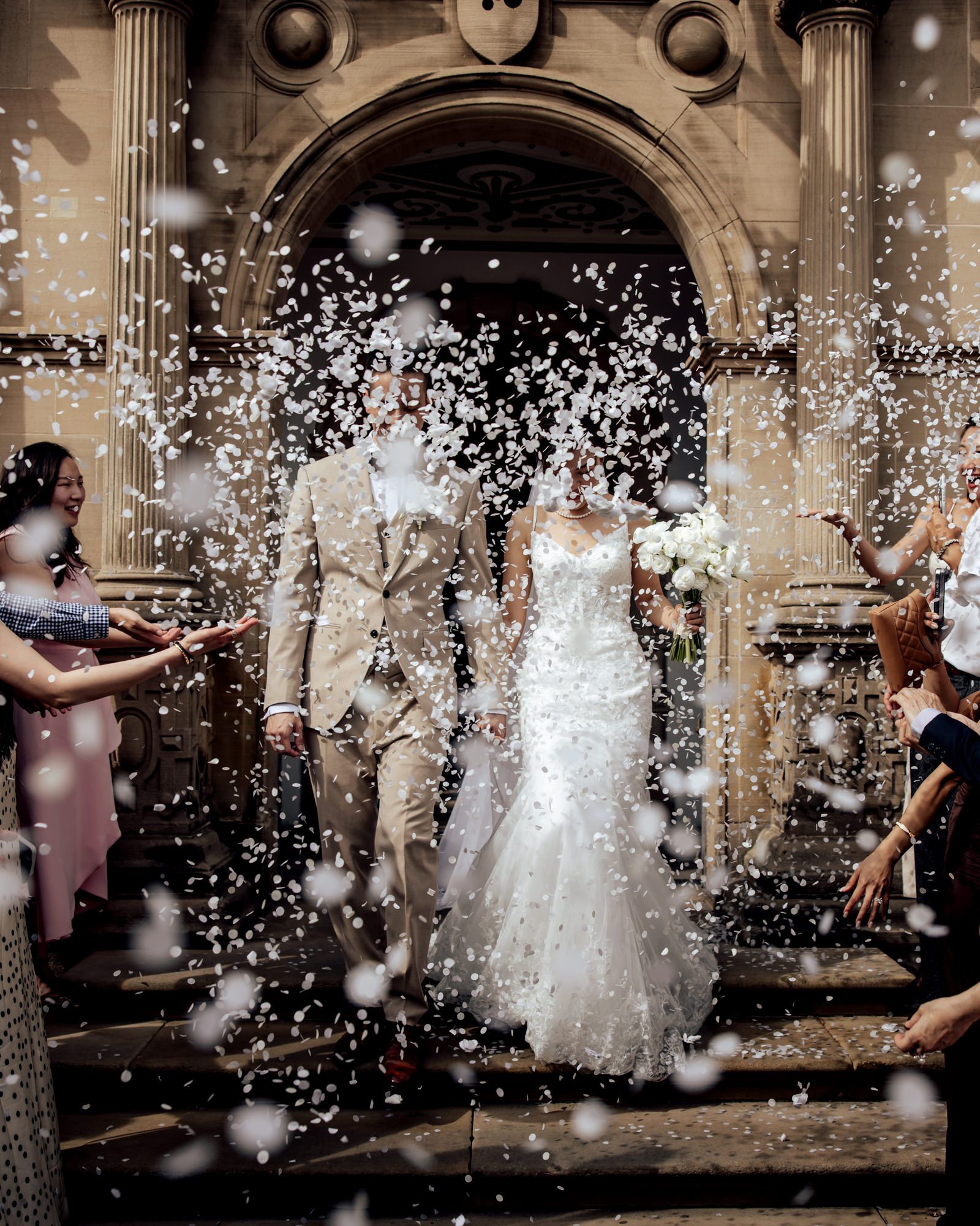 Newlyweds exit church in confetti shower, bride in white gown holding bouquet, groom in tan suit, guests celebrating.