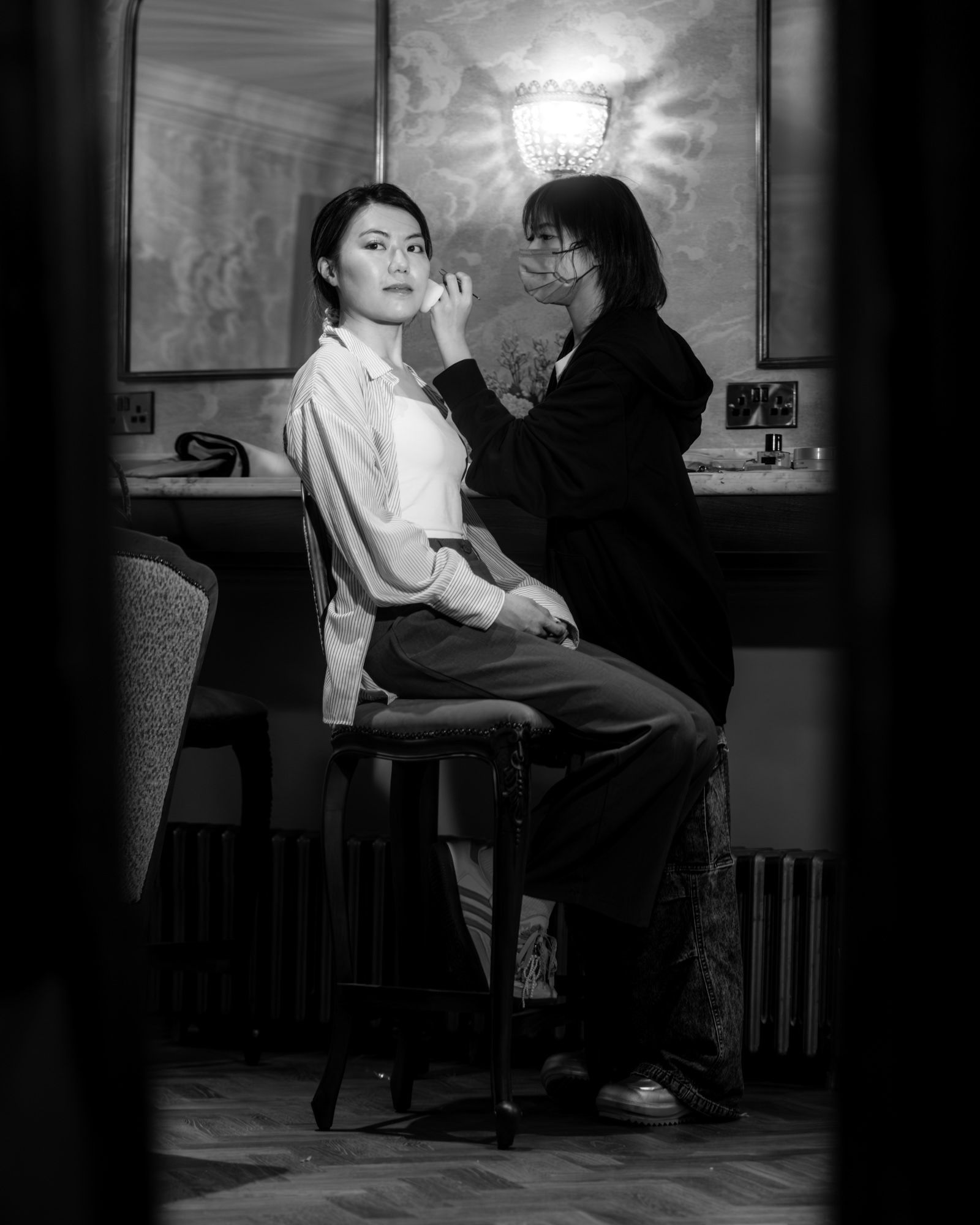 Black and white photo of a makeup artist applying makeup to a seated woman in an elegant room.