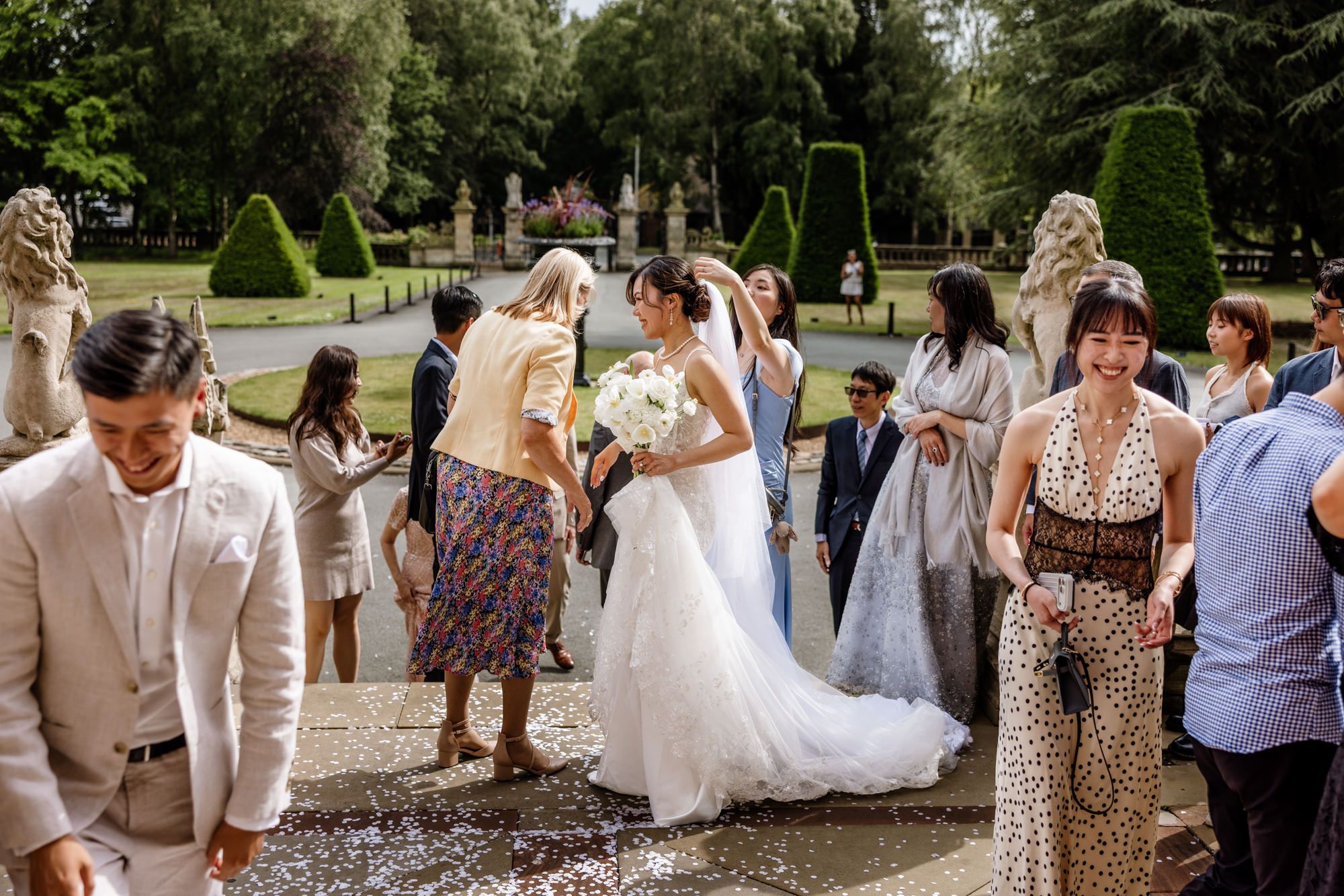 Bride in white gown walks through confetti shower among smiling guests in elegant garden wedding venue.