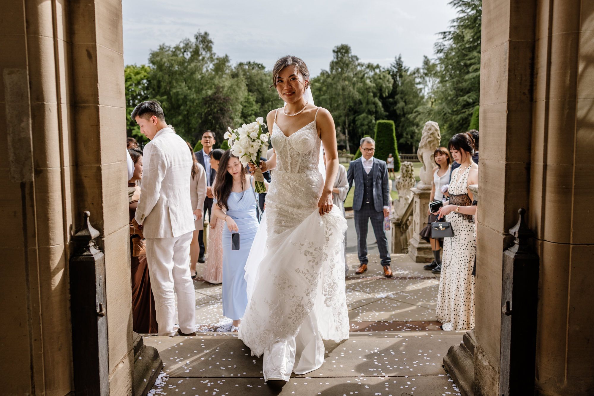 Bride in lace wedding gown walks through stone doorway at outdoor ceremony with guests and wedding party.