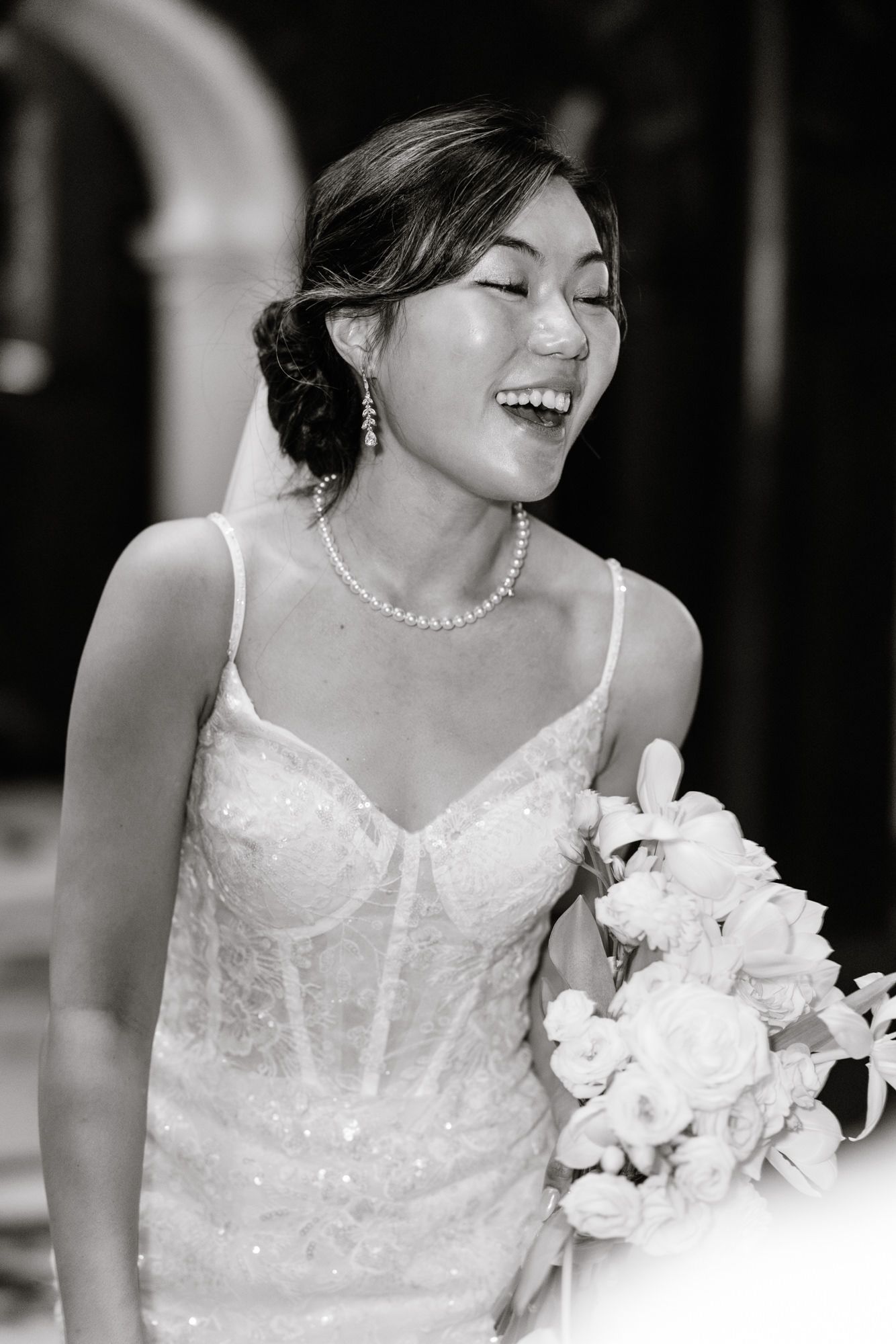 Joyful Asian bride laughing in a sequined white gown with pearl necklace and earrings, holding a white floral bouquet.
