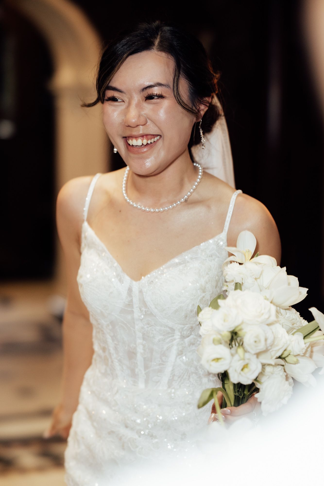 Smiling Asian bride in white sequin gown holding white bouquet, wearing pearl necklace and earrings with veil.
