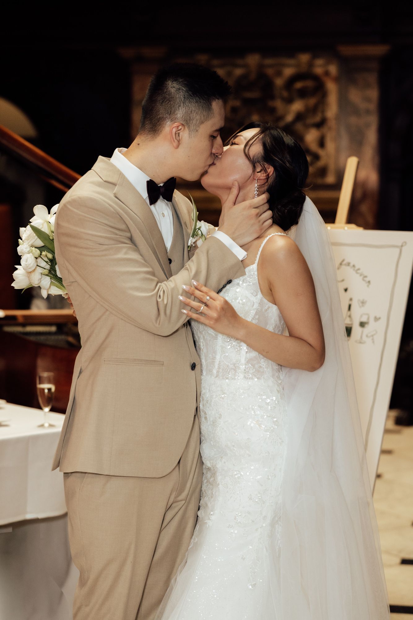 Newlyweds sharing a romantic first kiss, groom in beige suit and bride in lace wedding gown with veil.