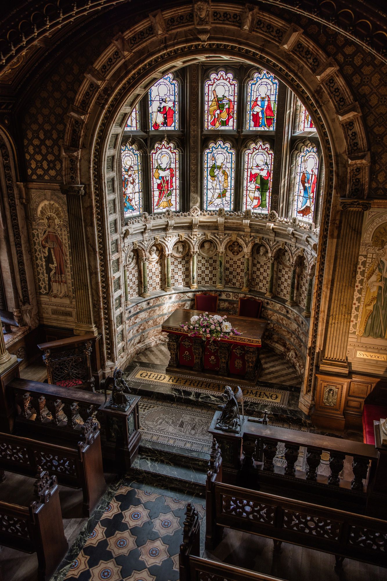 Interior of ornate church with stained glass windows, wooden pews, decorative arch, and floral altar arrangement.