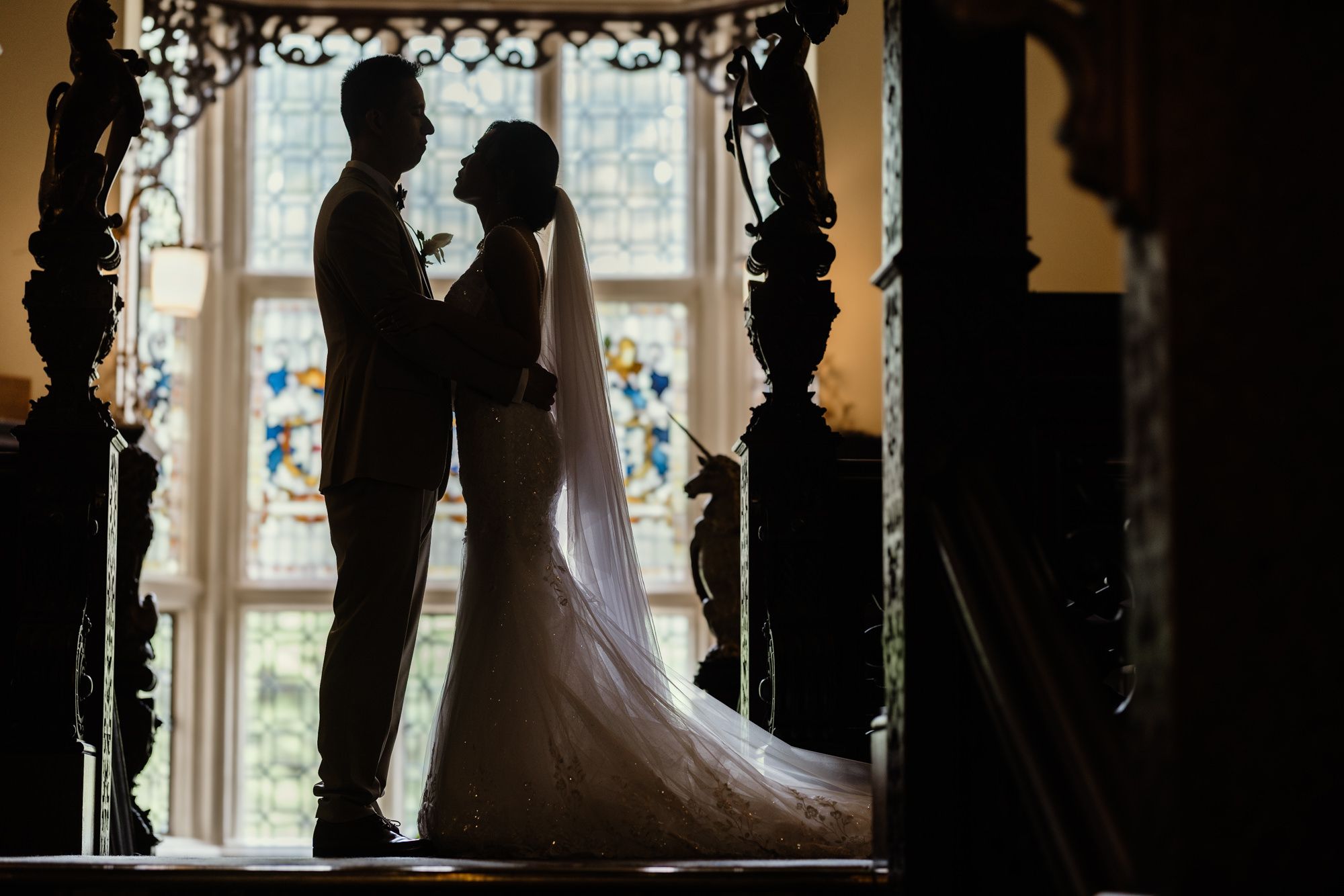 Silhouette of bride and groom standing before stained glass window in elegant church during romantic wedding ceremony.