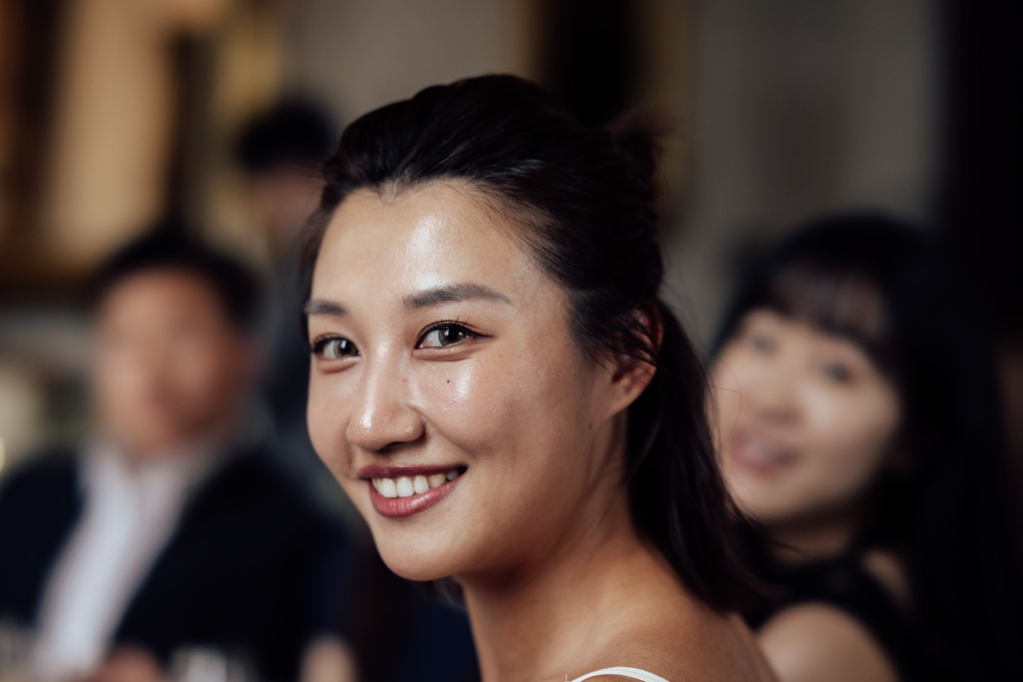 Smiling young Asian woman at a social gathering, with blurred guests in the background.