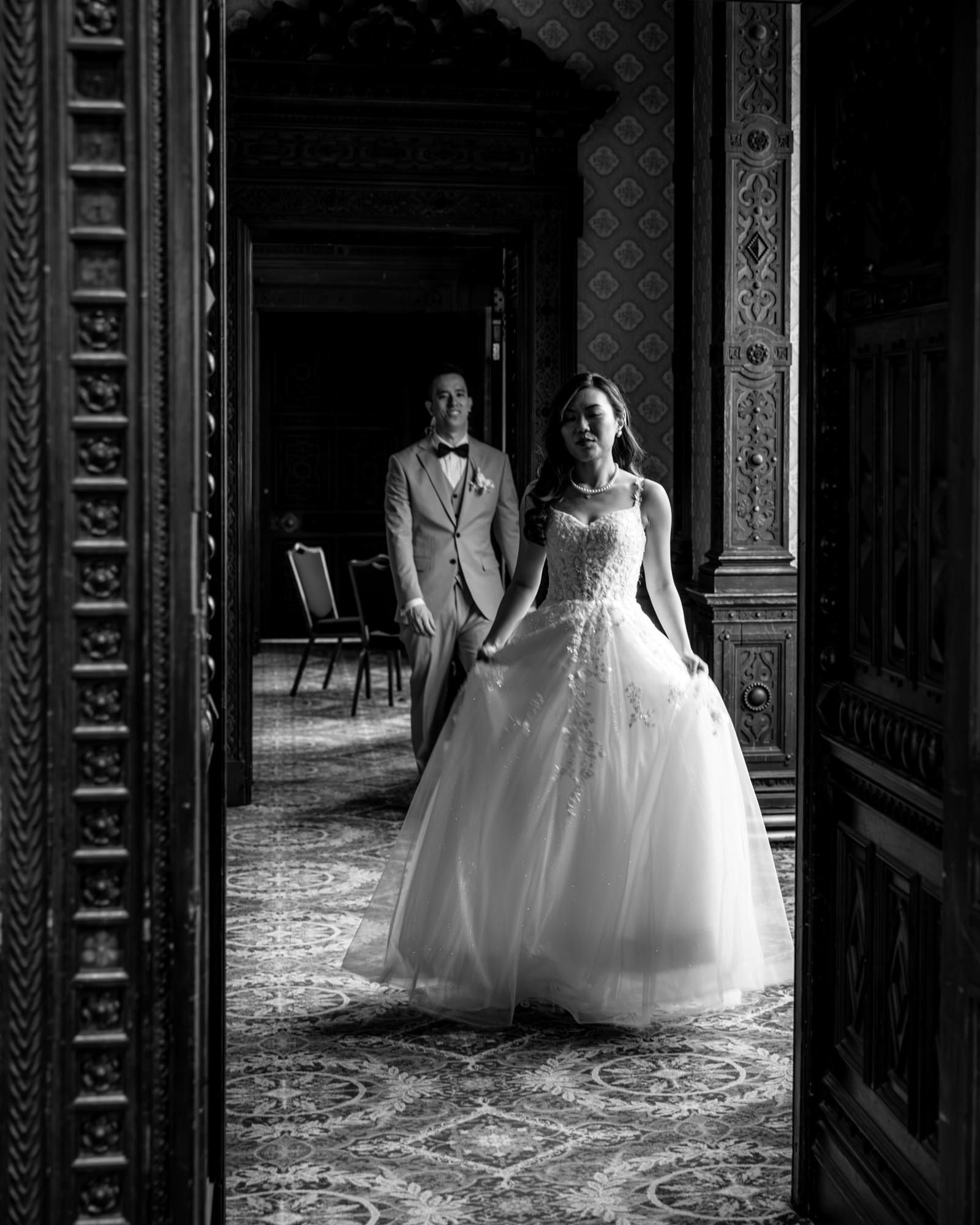 Black and white wedding photo of bride in ball gown and groom in suit walking through ornate doorway.