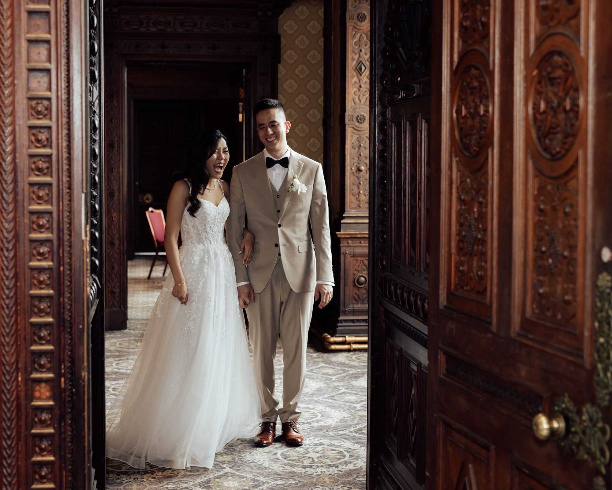 Bride in white tulle gown and groom in tan suit pose in ornate wooden doorway of elegant wedding venue.