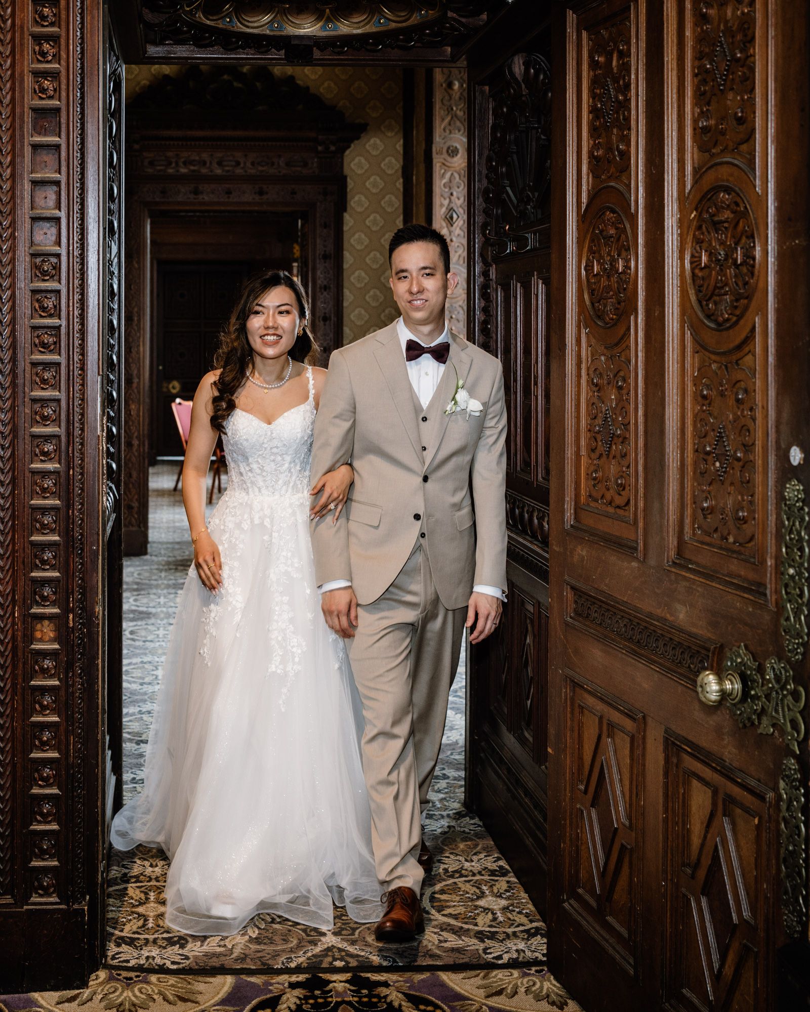 Bride in white lace gown and groom in tan suit walk through ornate wooden doors at elegant wedding venue.